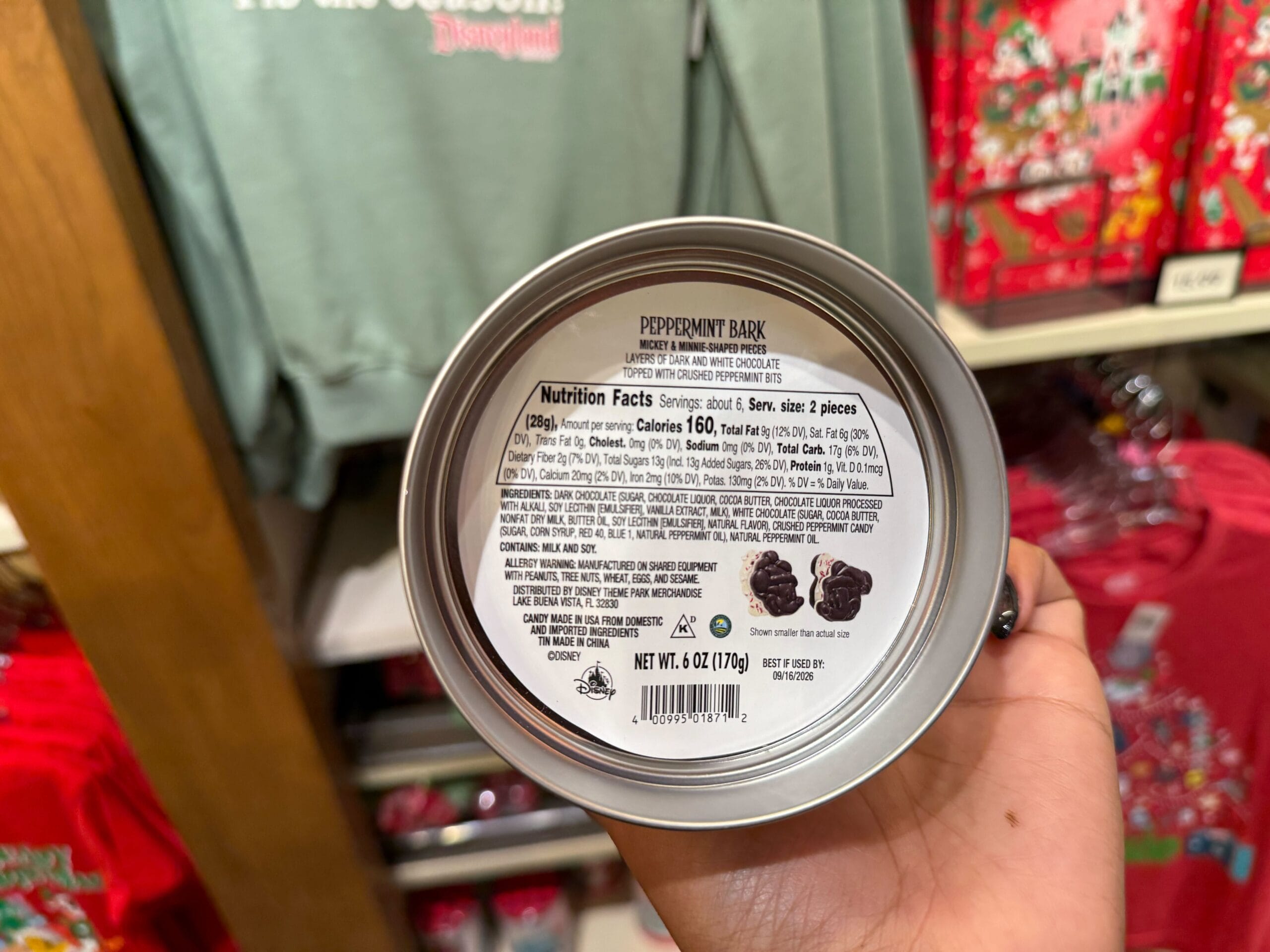 A hand holds a tin of peppermint bark from the Peppermint Collection in a shop filled with Disneyland holiday merchandise.