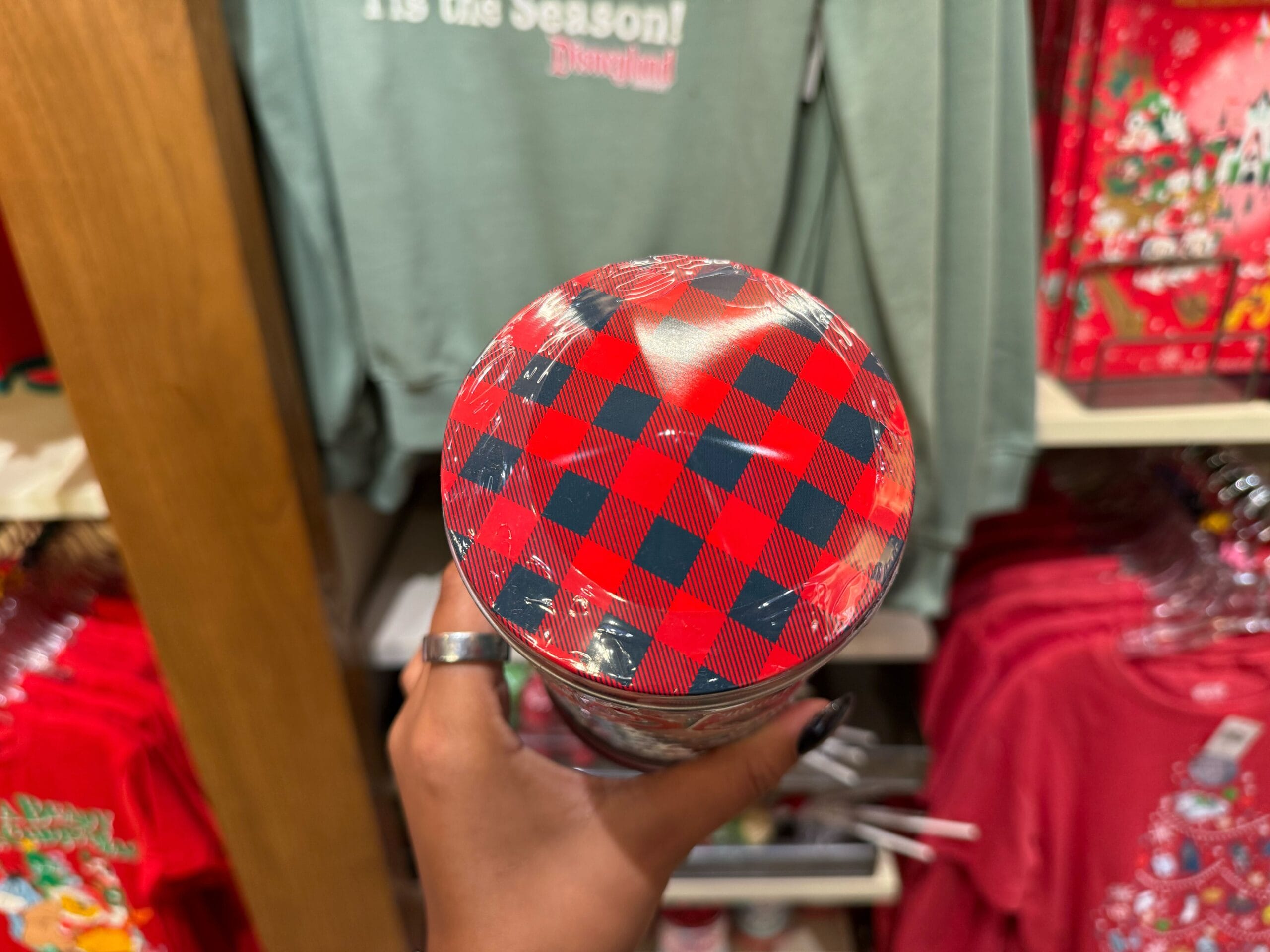 A hand holds a cup with a plaid lid inside a Disney park shop, surrounded by Peppermint Collection holiday clothing displays.