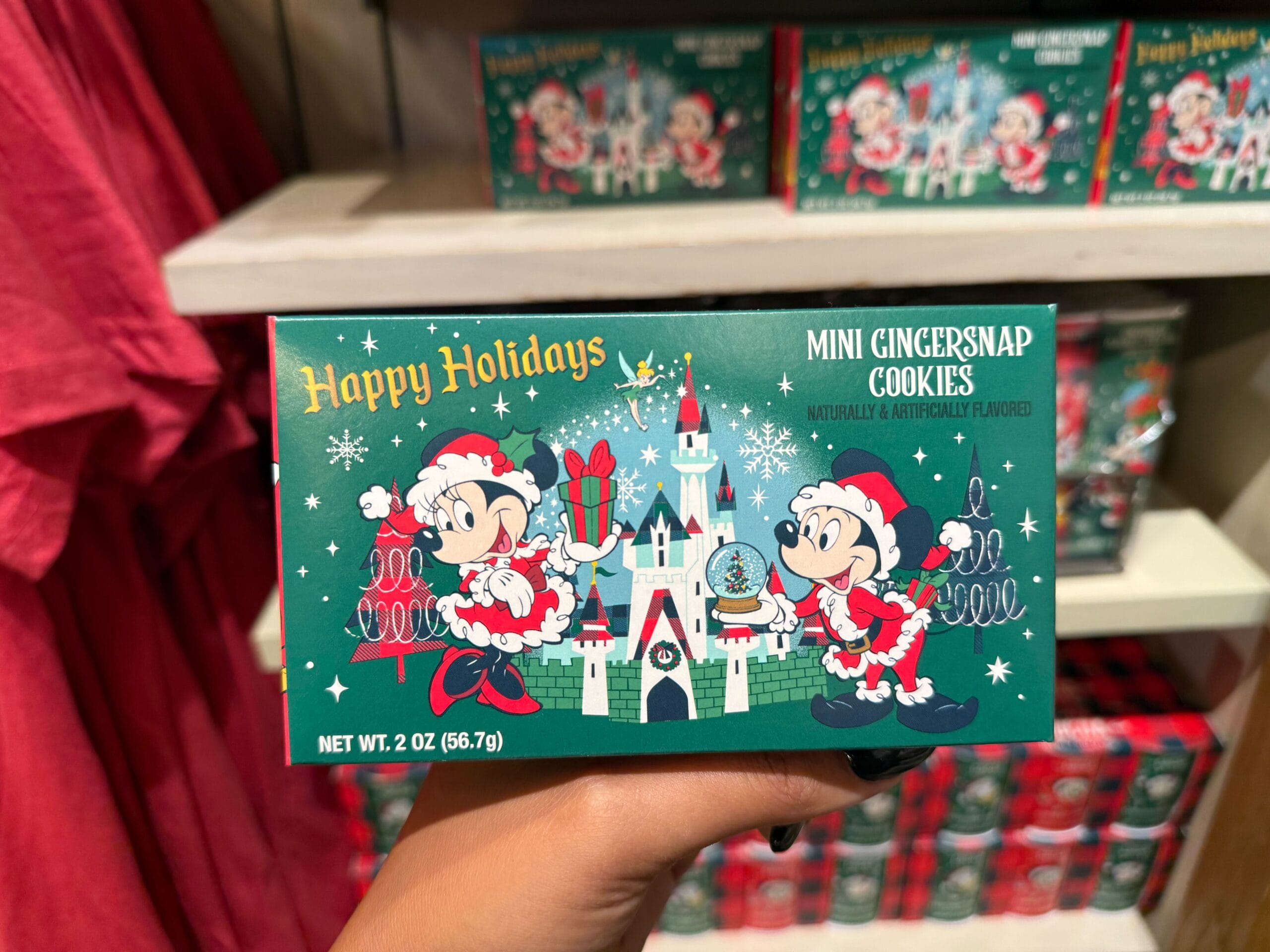 A hand holds a green box of Disneyland Christmas mini gingersnap cookies near a castle resembling Sleeping Beauty Castle in Anaheim.