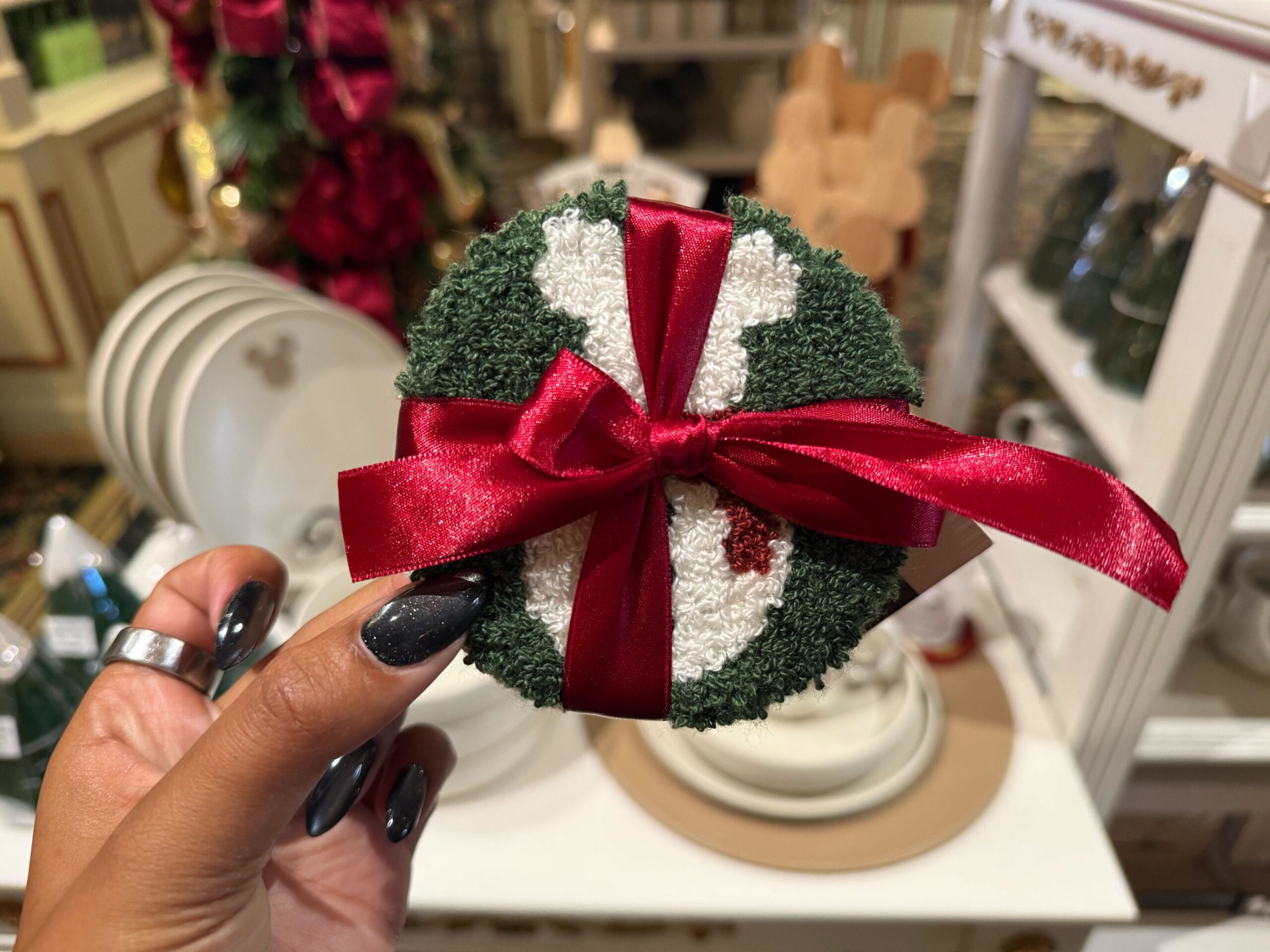 A hand displays a green and white Disneyland Resort ornament with a red ribbon, surrounded by festive holiday park merchandise.
