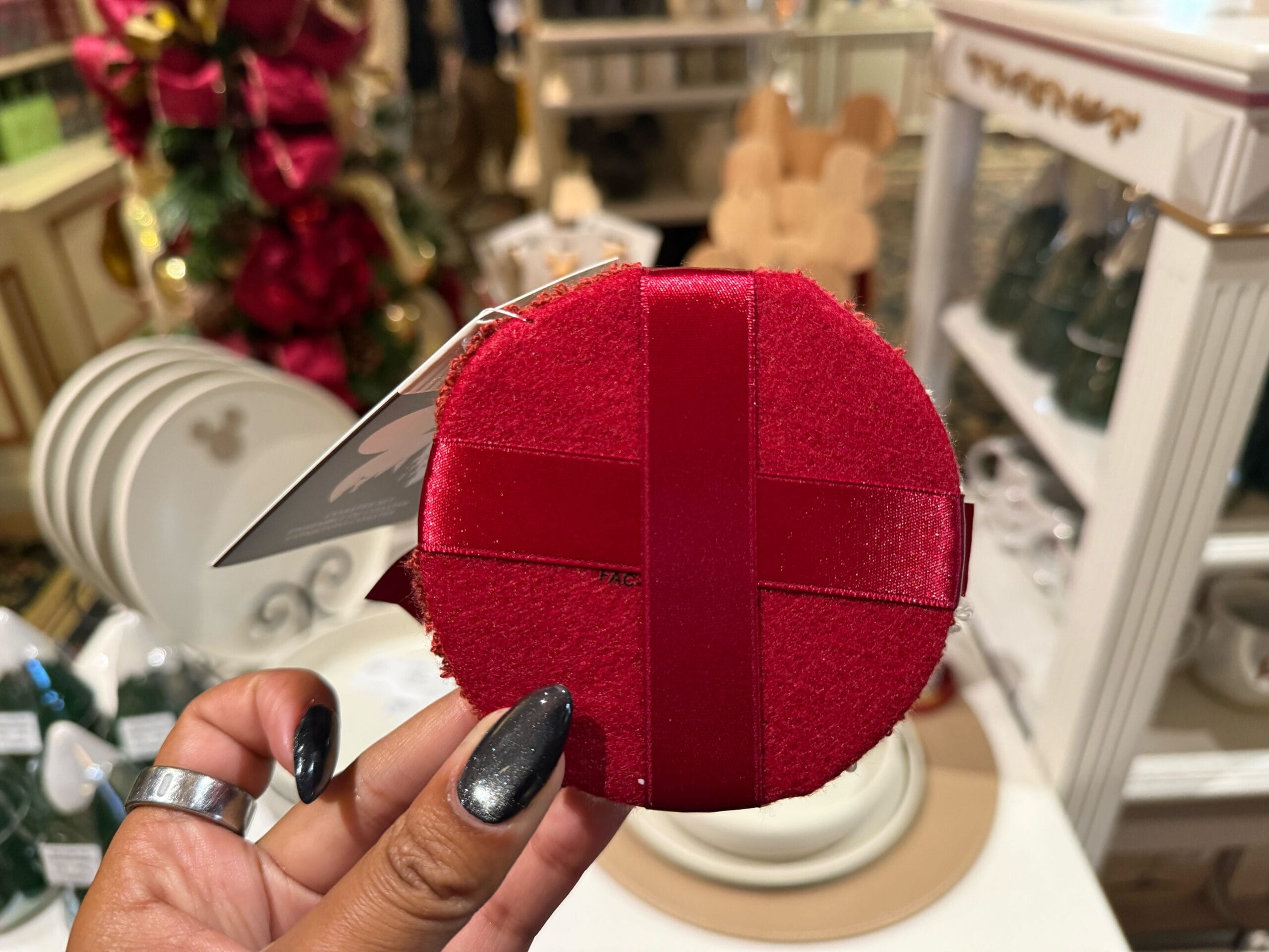 A hand holds a red velvet Disney Christmas ornament with a ribbon in a shop filled with festive holiday decor and themed dinnerware.
