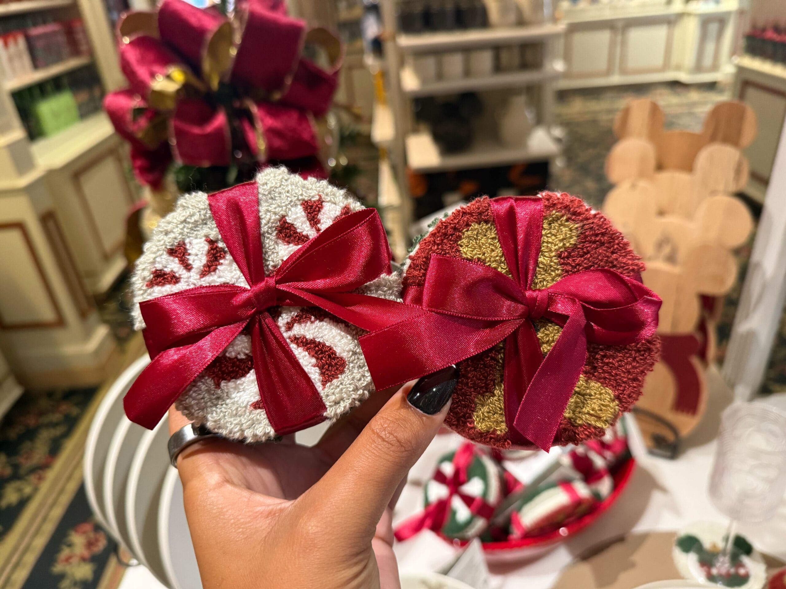 A hand holds two round Disney Christmas ornaments with red ribbon bows—new arrivals among festive park merchandise this season.