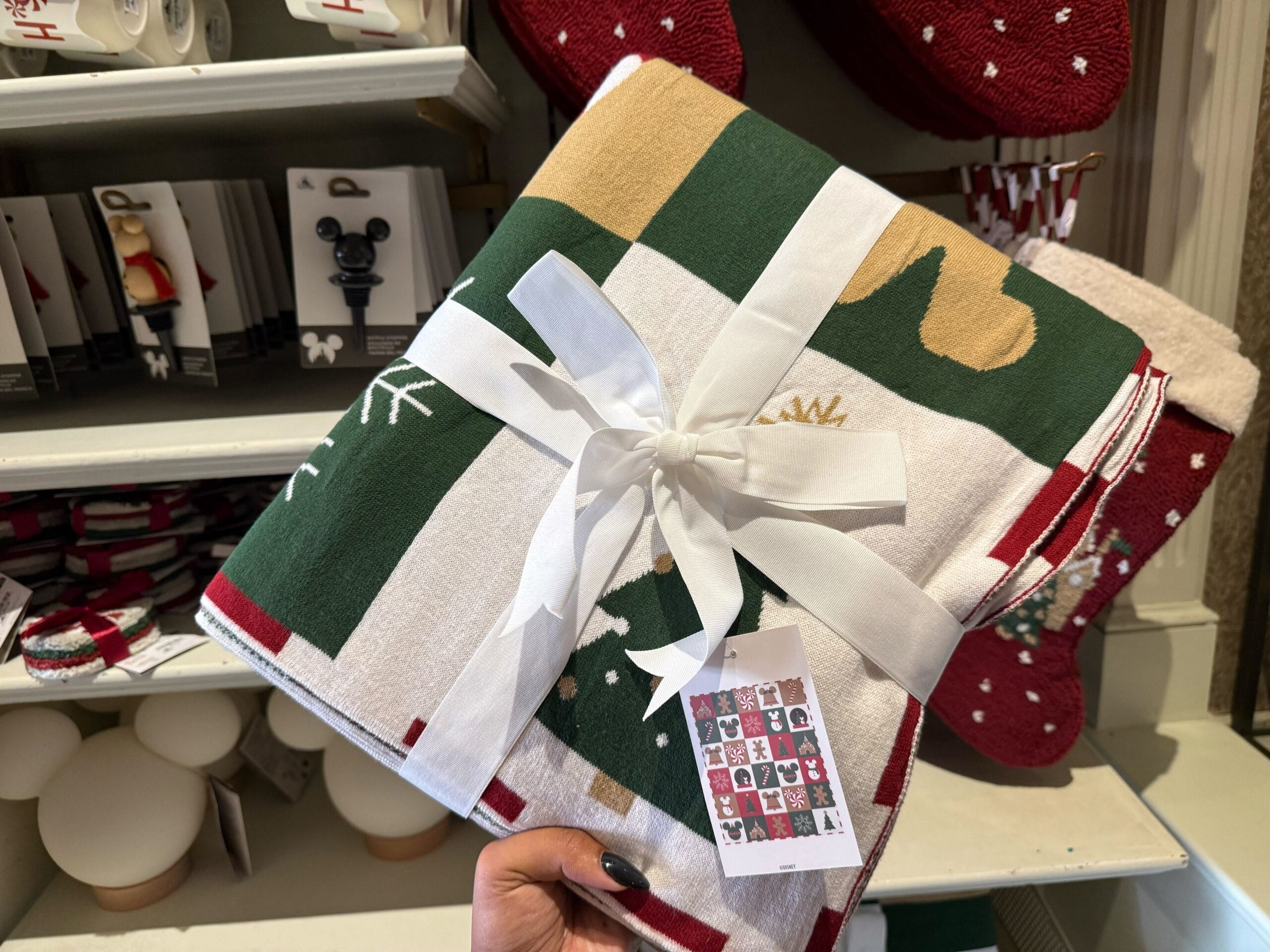 A hand displays a folded, ribbon-tied Christmas blanket from Disneyland Resort’s latest holiday merchandise lineup. No castle visible.