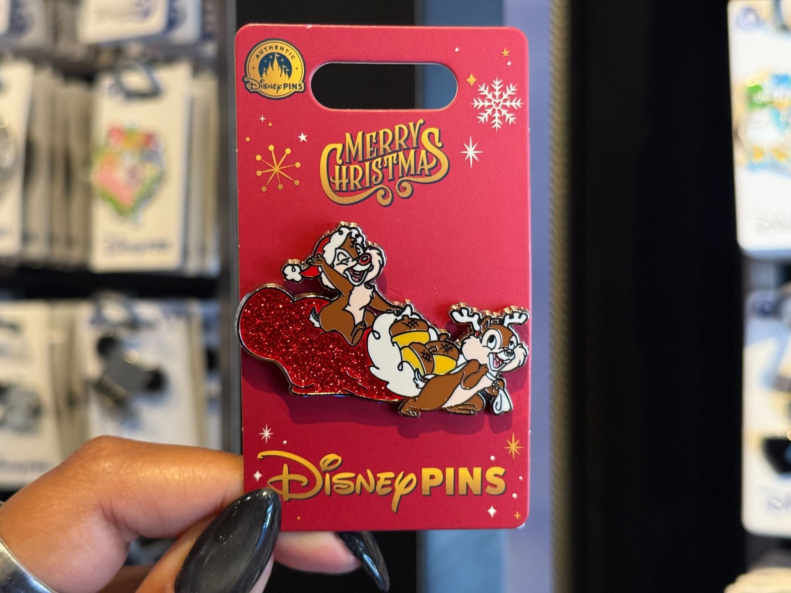 A hand holds Disneyland Resort Christmas merch: a Disney pin with Chip and Dale on a red sled—no castle, so no park comparison needed.