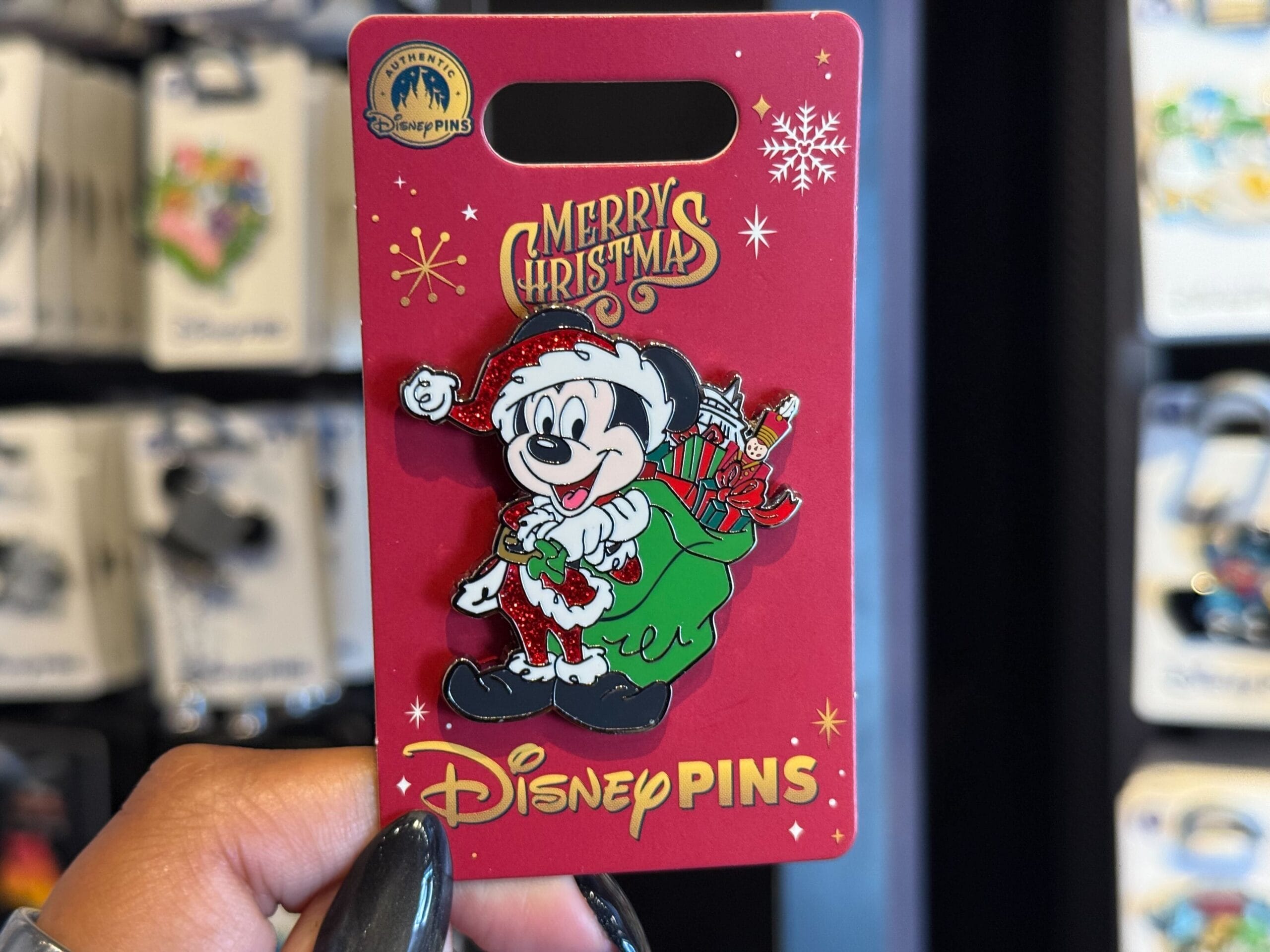 A hand holds Disneyland Resort holiday merchandise: a Mickey Mouse Santa pin with green gift sack, red card—no castle in view.