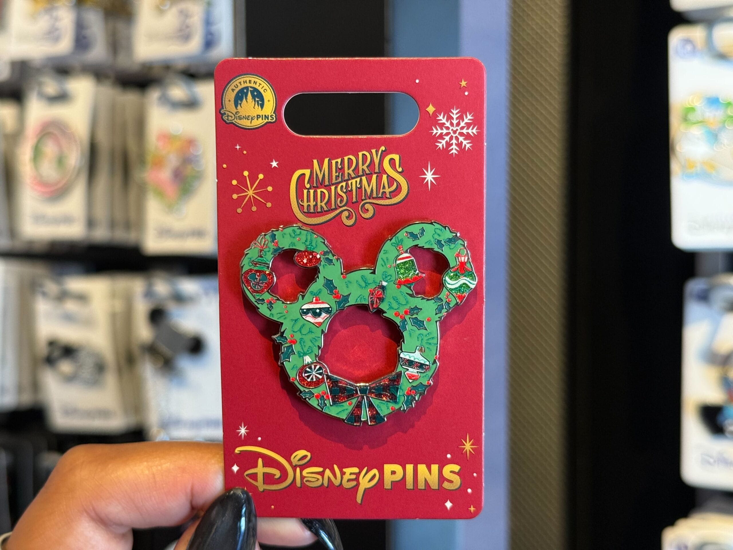 A hand holds a Disneyland Resort pin shaped like a Mickey wreath on a red "Merry Christmas" card, evoking classic park holiday cheer.