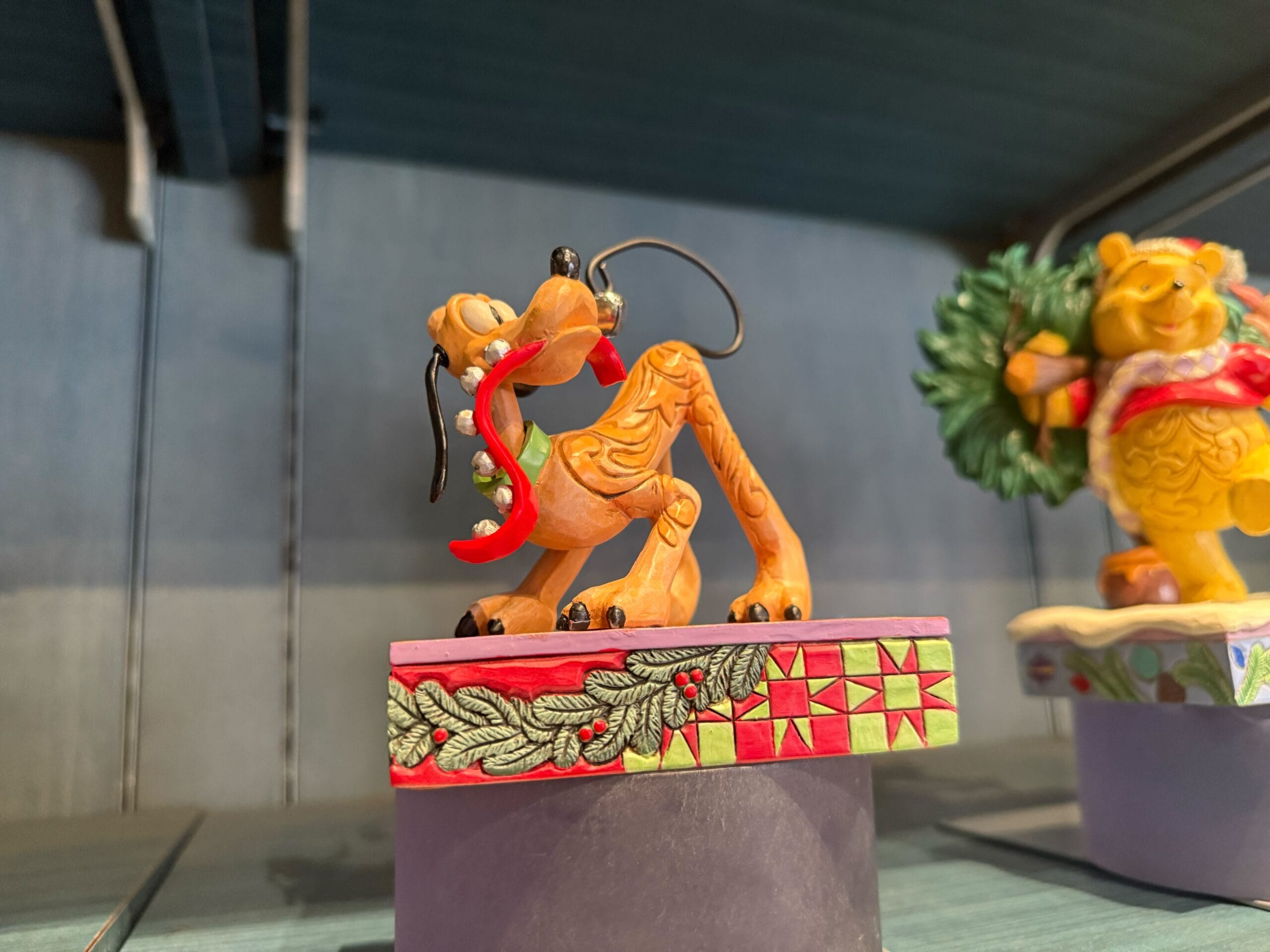 A festive Pluto figurine with a red scarf stands on a Disney Christmas-themed base, reminiscent of holiday displays in Disney Parks.