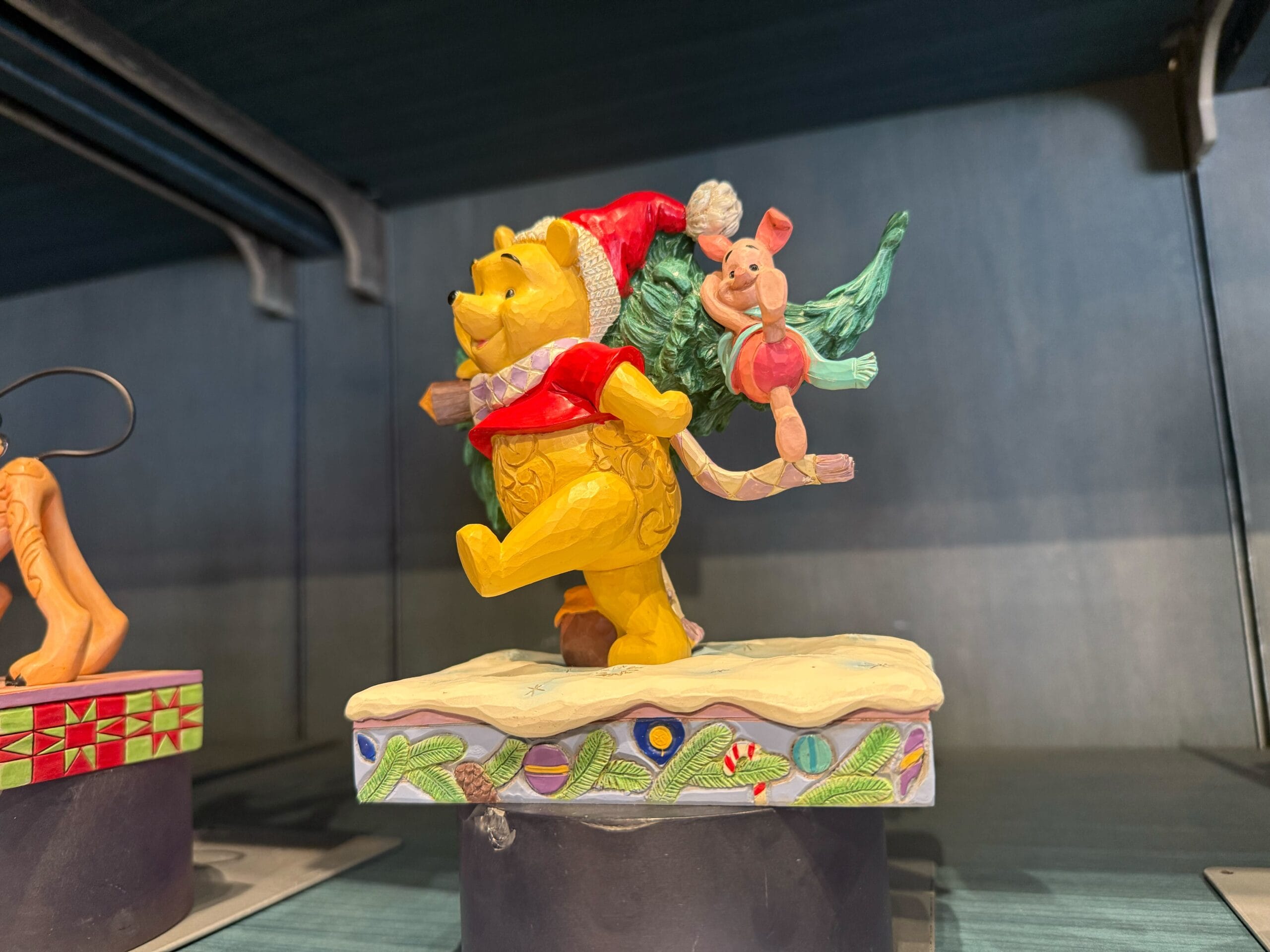 Winnie the Pooh in a Santa hat carries a Christmas tree with Piglet—festive Disneyland Resort merch, no castle like Disneyland’s nearby.