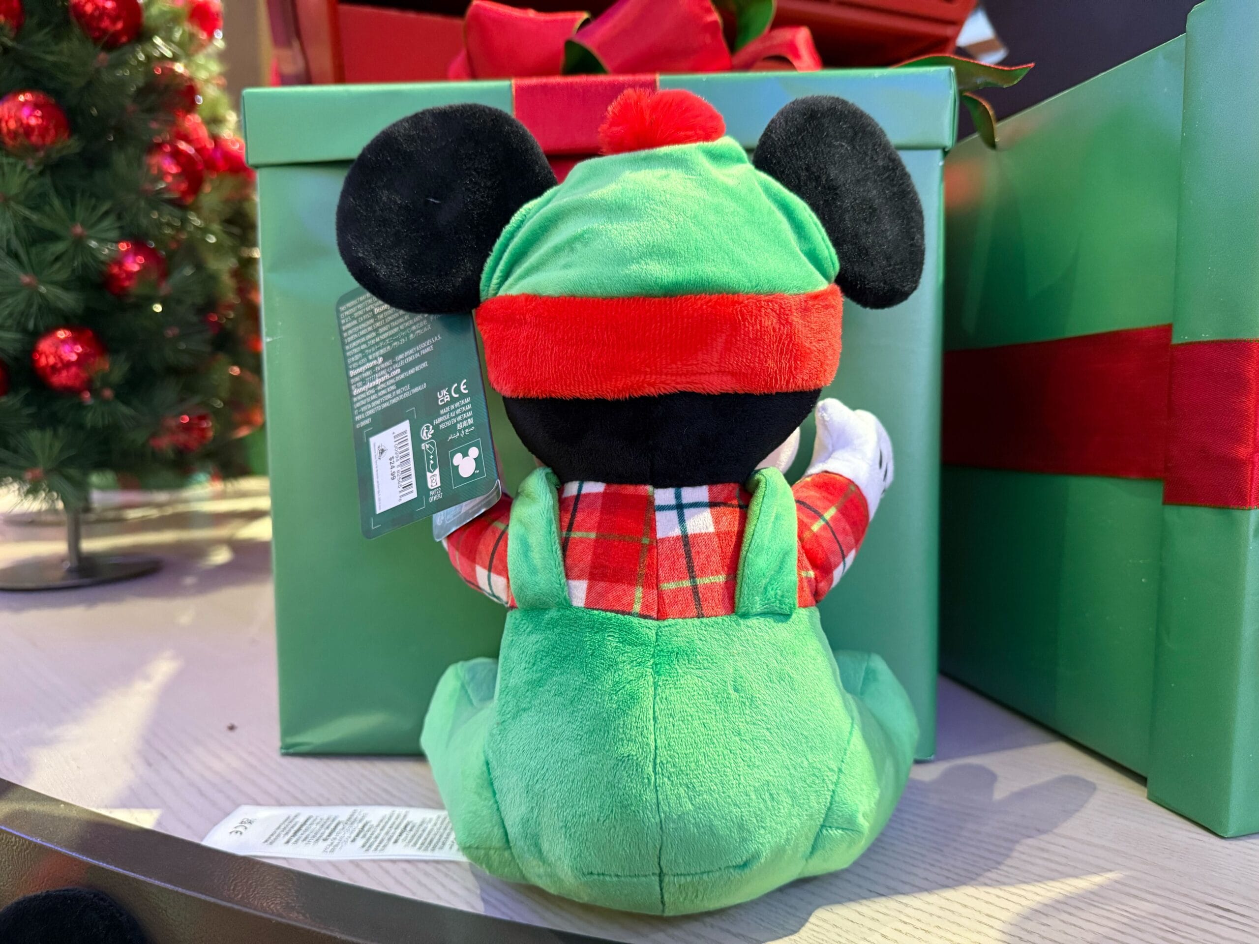 Festive plush in green overalls and red hat sits by a green gift box, featured in Disneyland Resort’s Christmas merchandise display.