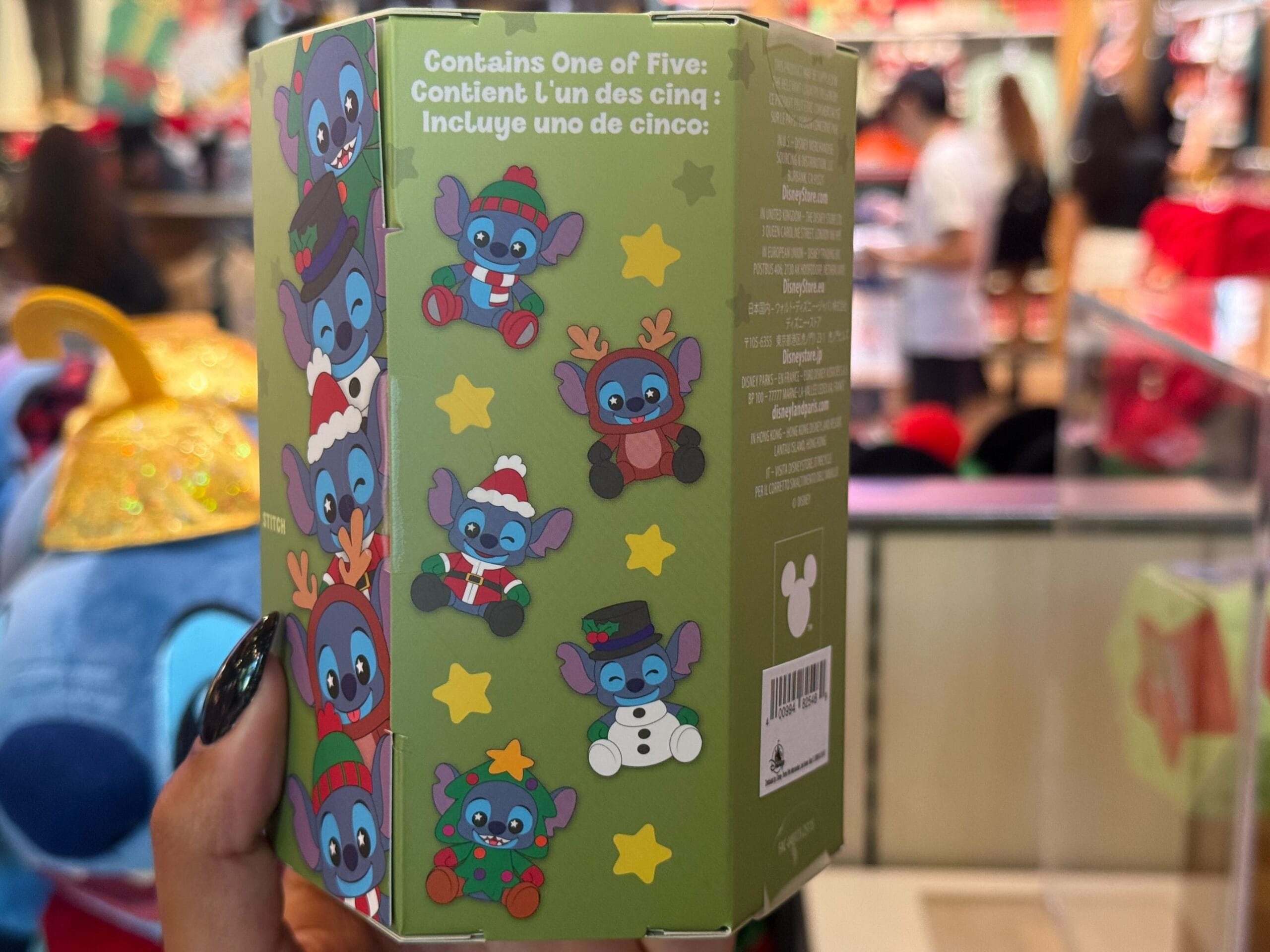 A hand holds a green Christmas Stitch box at Disneyland Resort, with stars—unlike the castle seen at Walt Disney World.