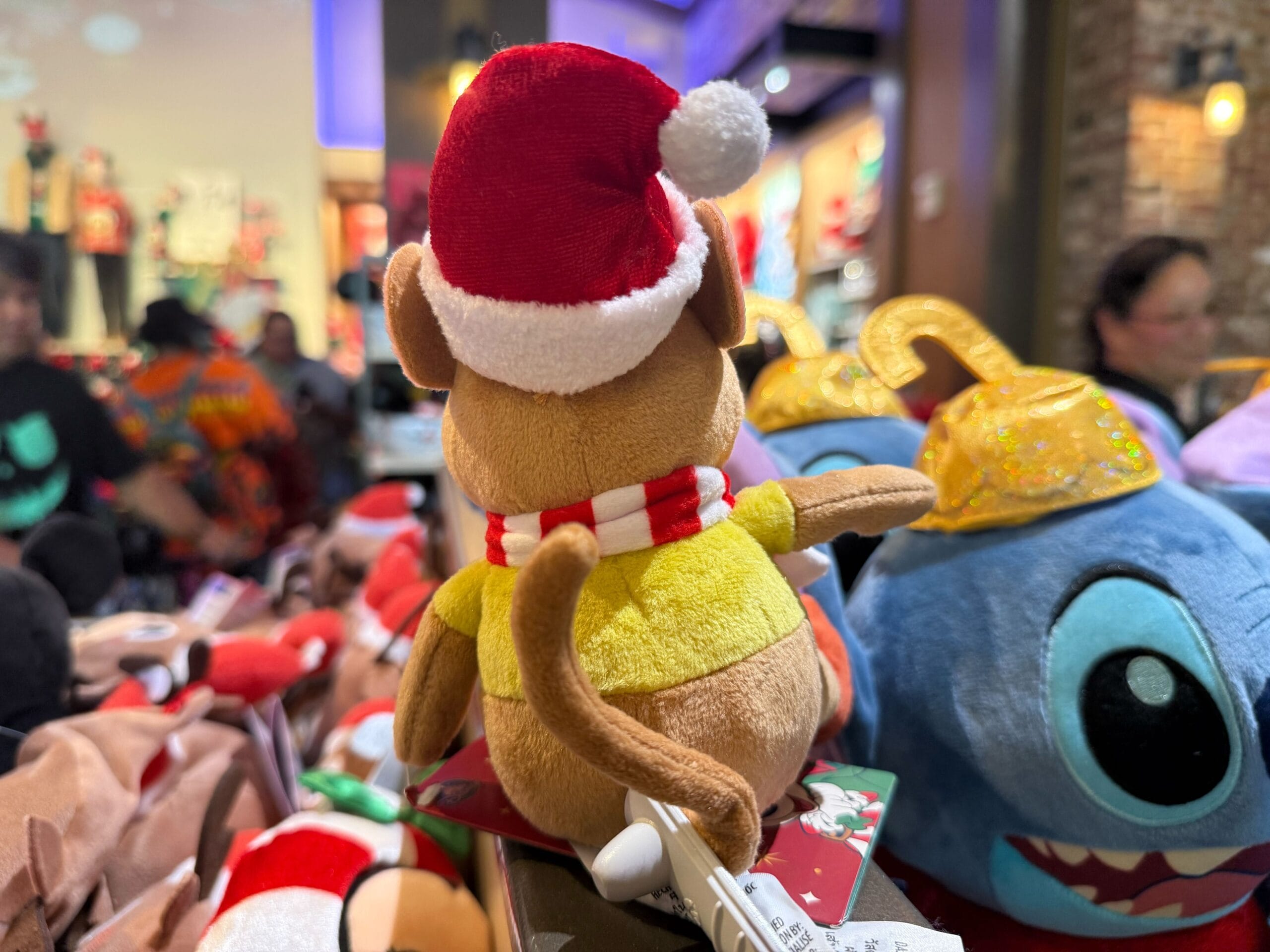 A plush monkey in a Santa hat and scarf faces away, surrounded by Disney holiday merchandise in a park store, festive and cheerful.