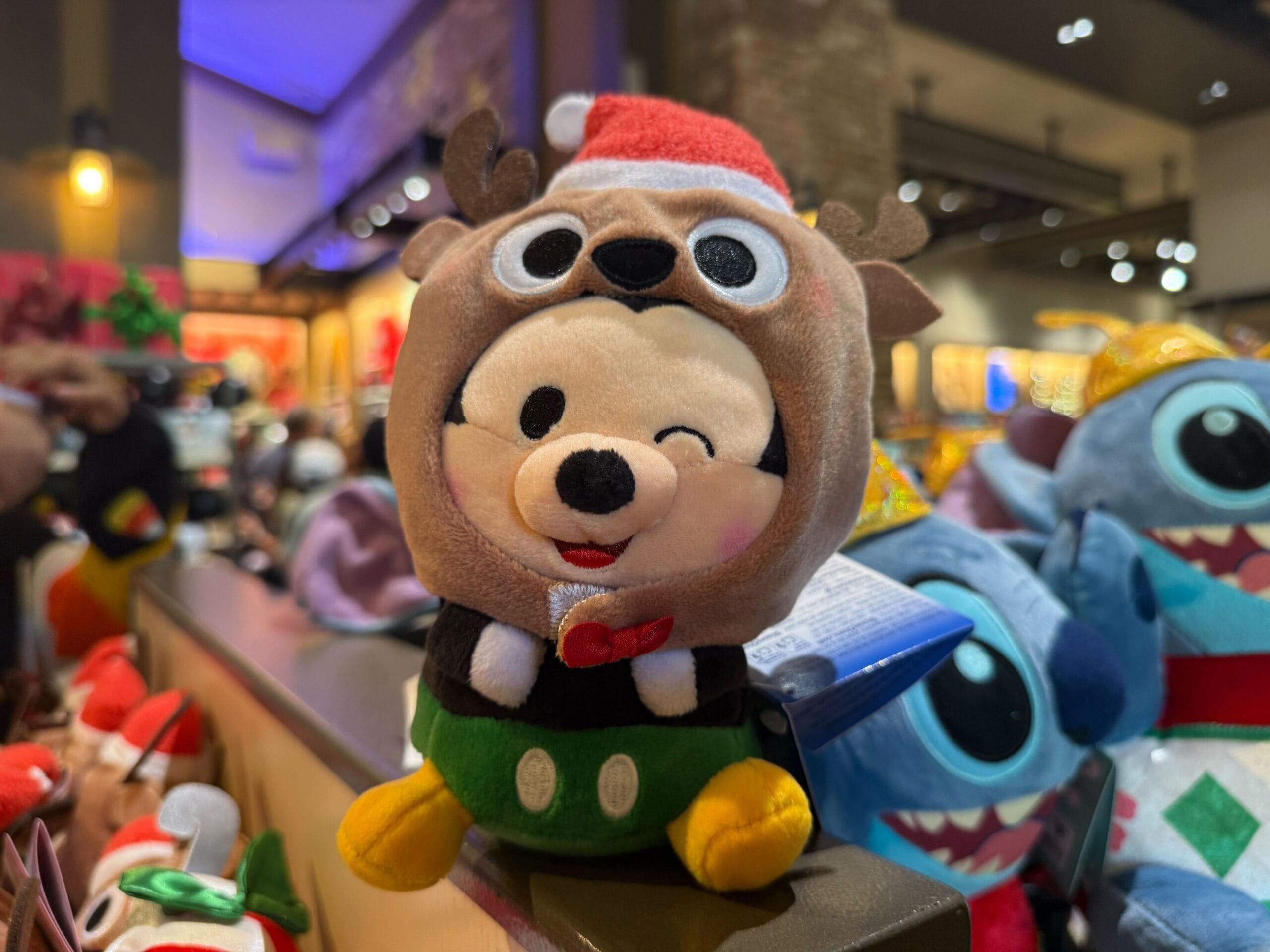A winking plush Mickey Mouse in a reindeer hat sits among festive Christmas goods at Disneyland Resort, with no castle in view.