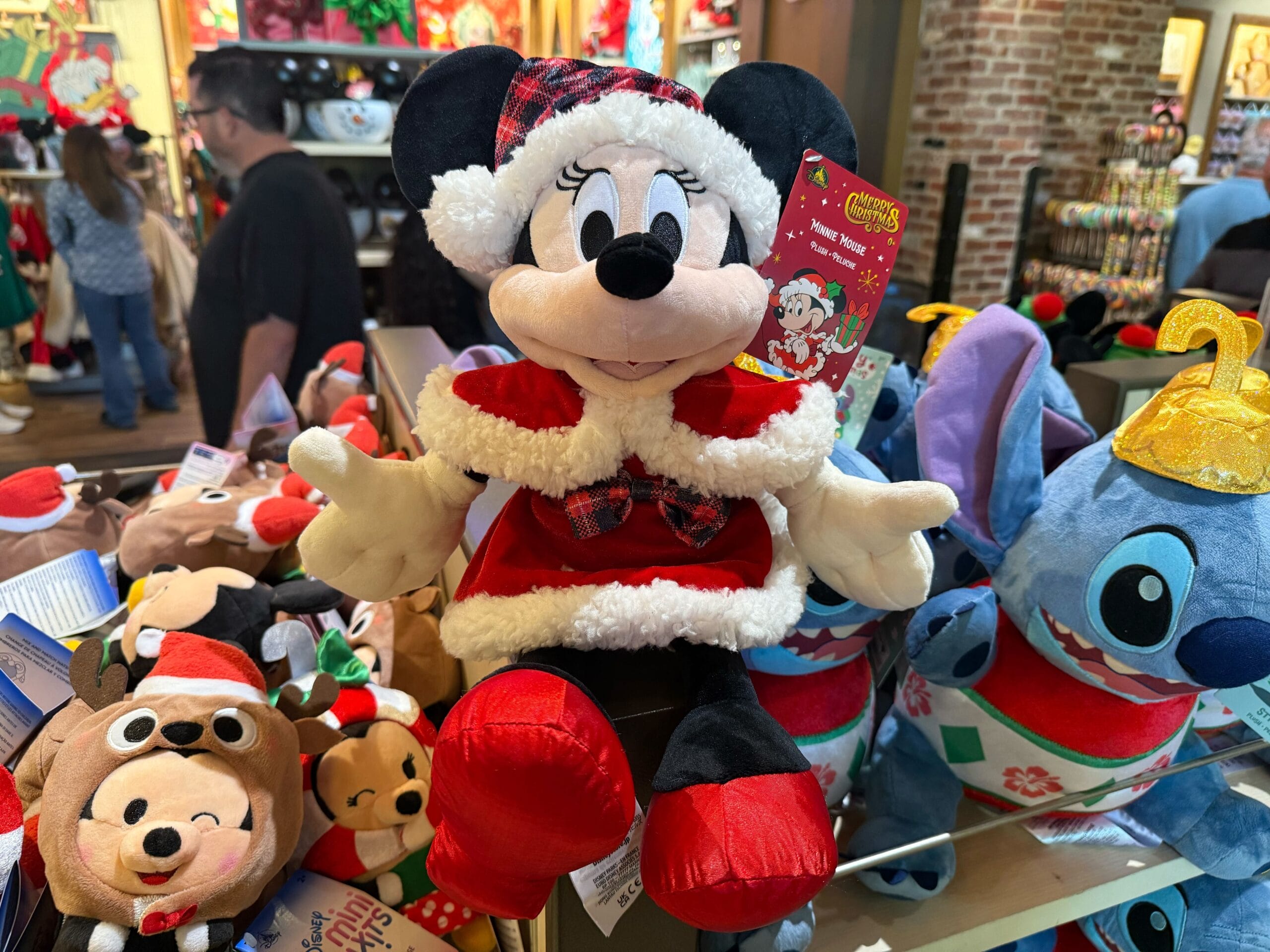 A Minnie Mouse plush in a red plaid Christmas outfit stands among holiday New Arrivals at Disneyland Resort, near Sleeping Beauty Castle.