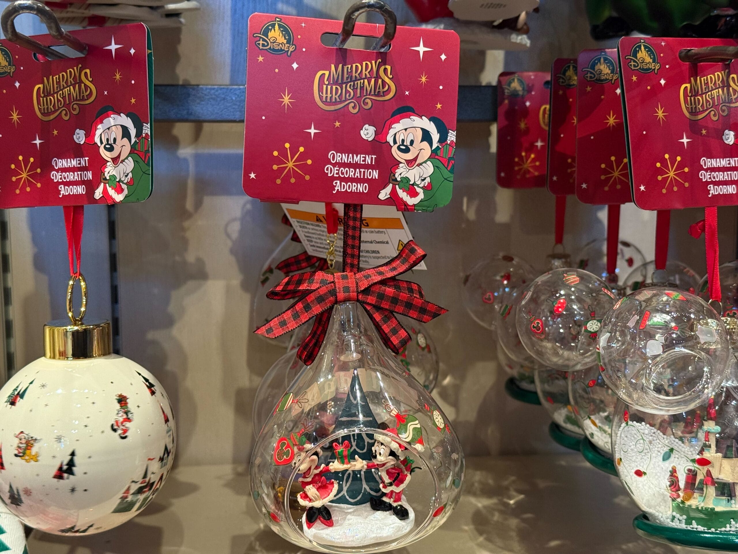 Clear glass ornament from Disneyland Resort featuring Minnie Mouse, plaid bow, snow, and red packaging on a park store shelf.
