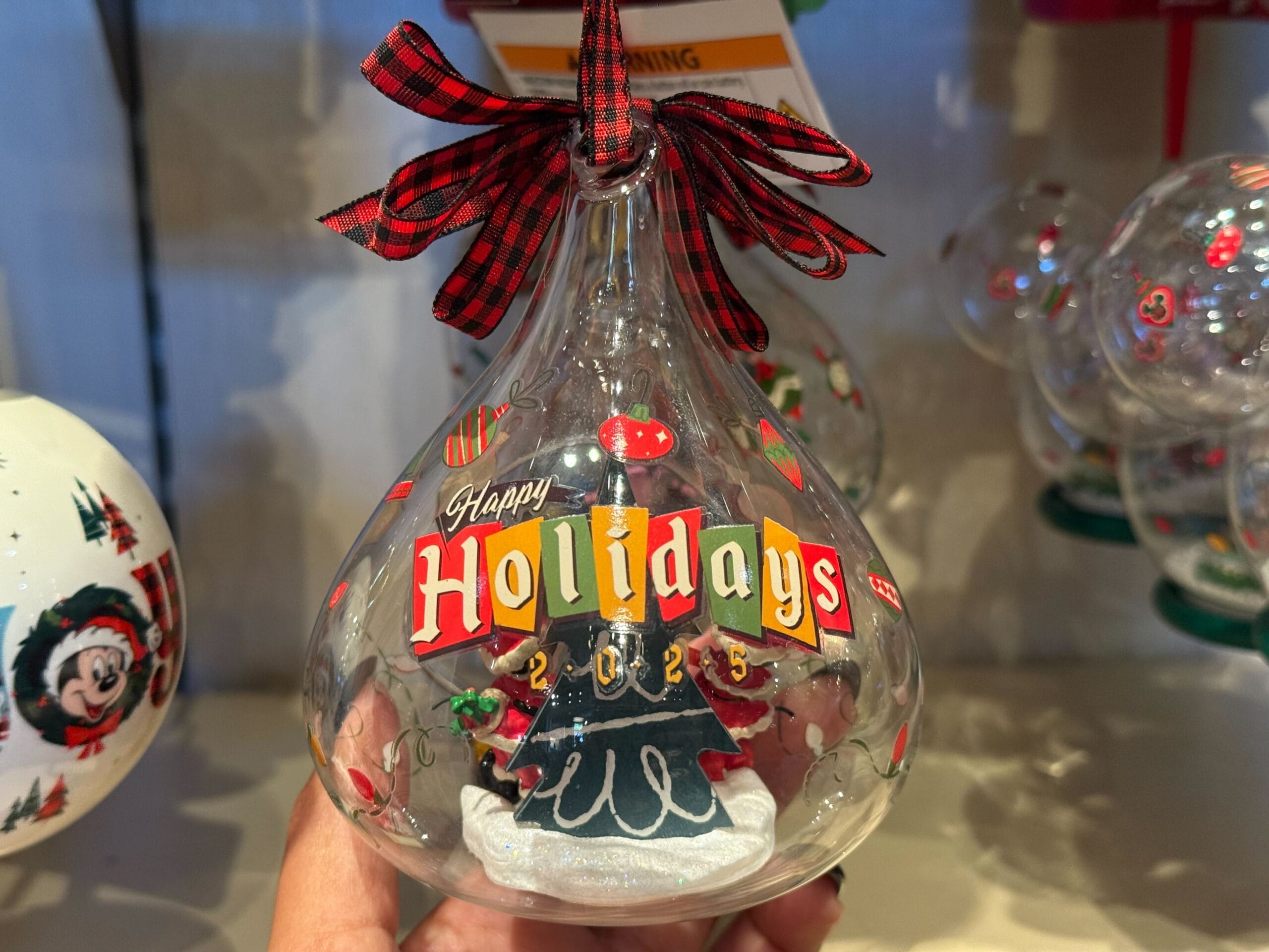 Glass ornament with a Christmas tree, "Happy Holidays 2025" text, and red plaid ribbon—iconic Disneyland Resort holiday style.