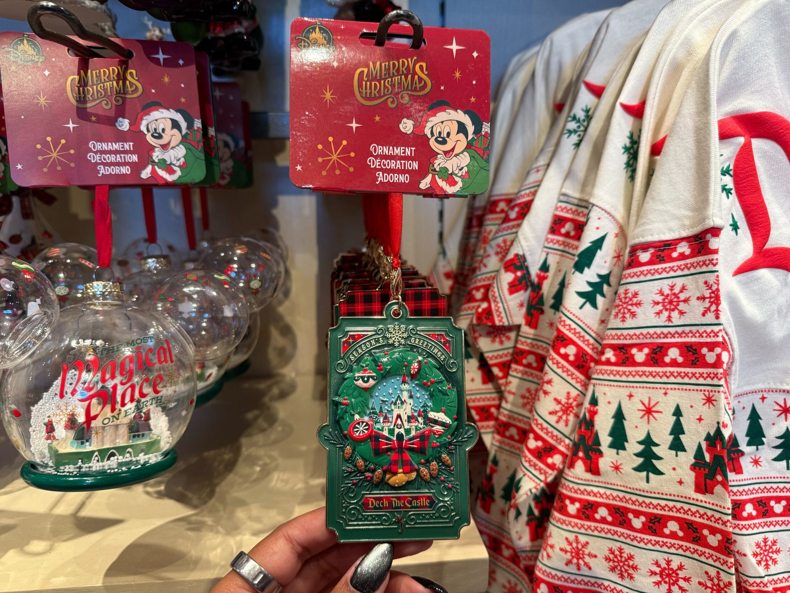 Hand holding a "Deck the Castle" ornament, festive shirts, and Mickey ear decor—Disneyland Resort merch with Sleeping Beauty Castle flair.