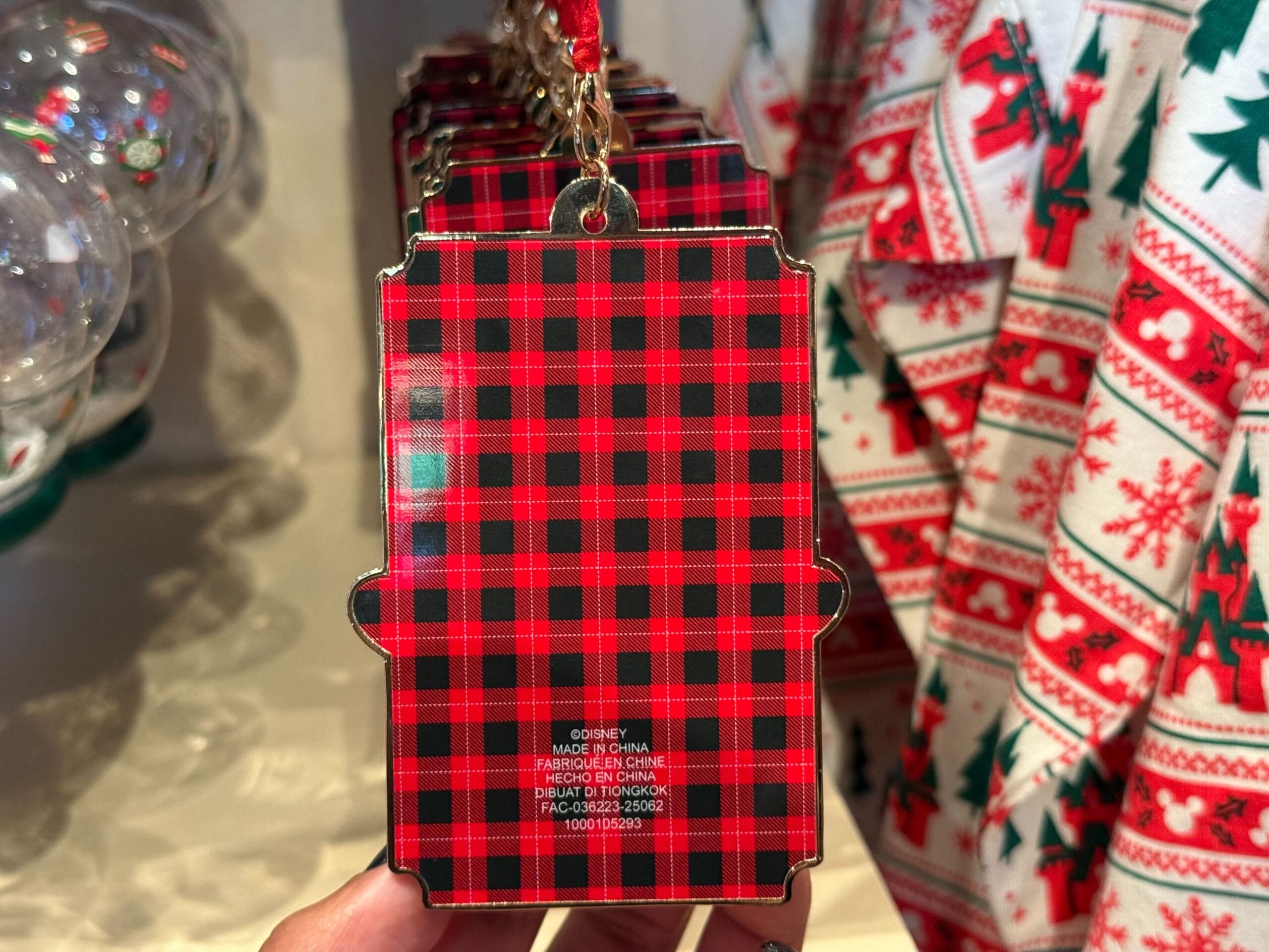 A hand holds a red and black plaid ornament with product details; Disney Park Christmas merchandise glows in a festive background.