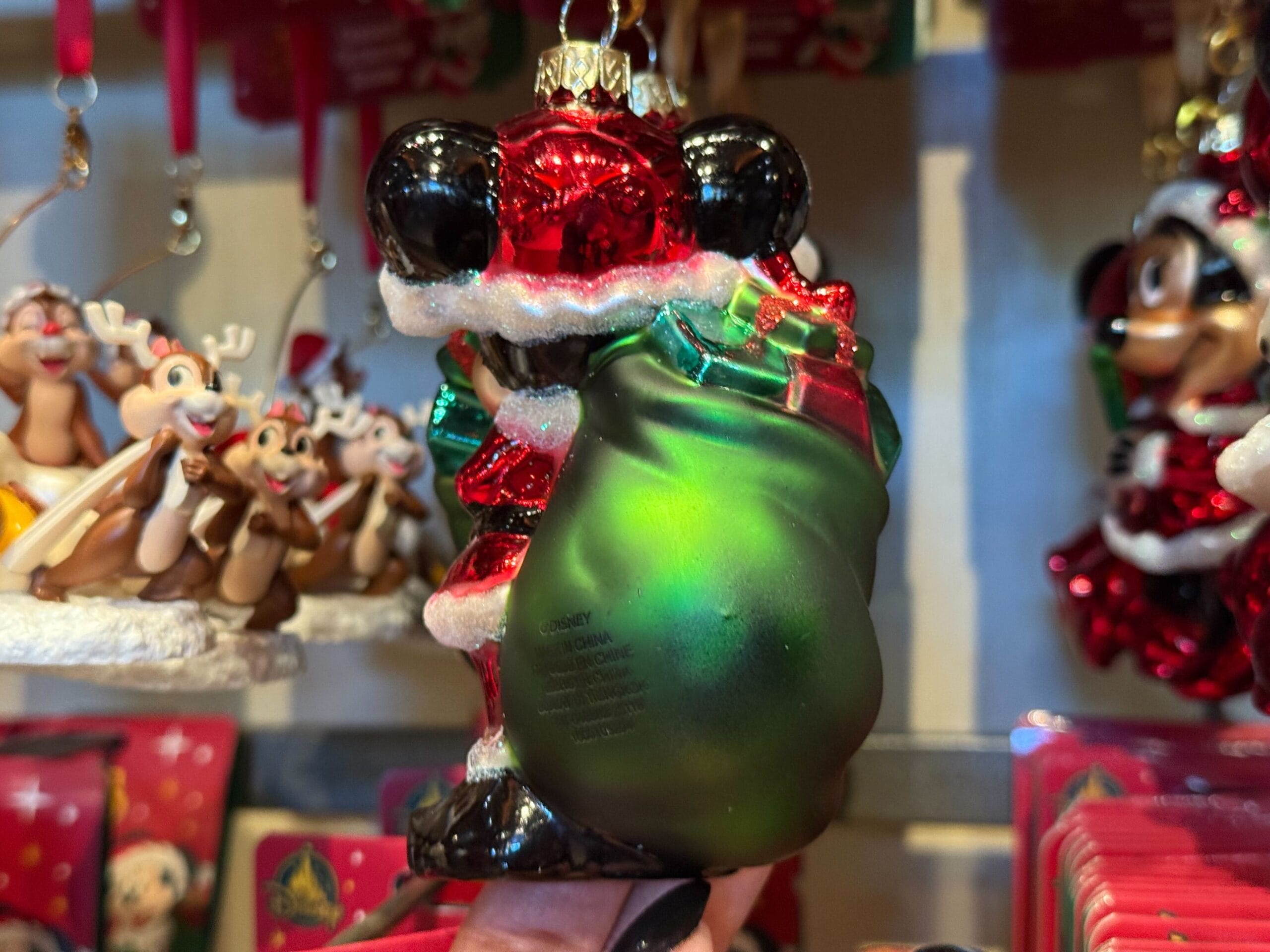A hand holds a Santa Mickey ornament from Disneyland Resort, with a green sack, surrounded by festive Christmas Disney merch.