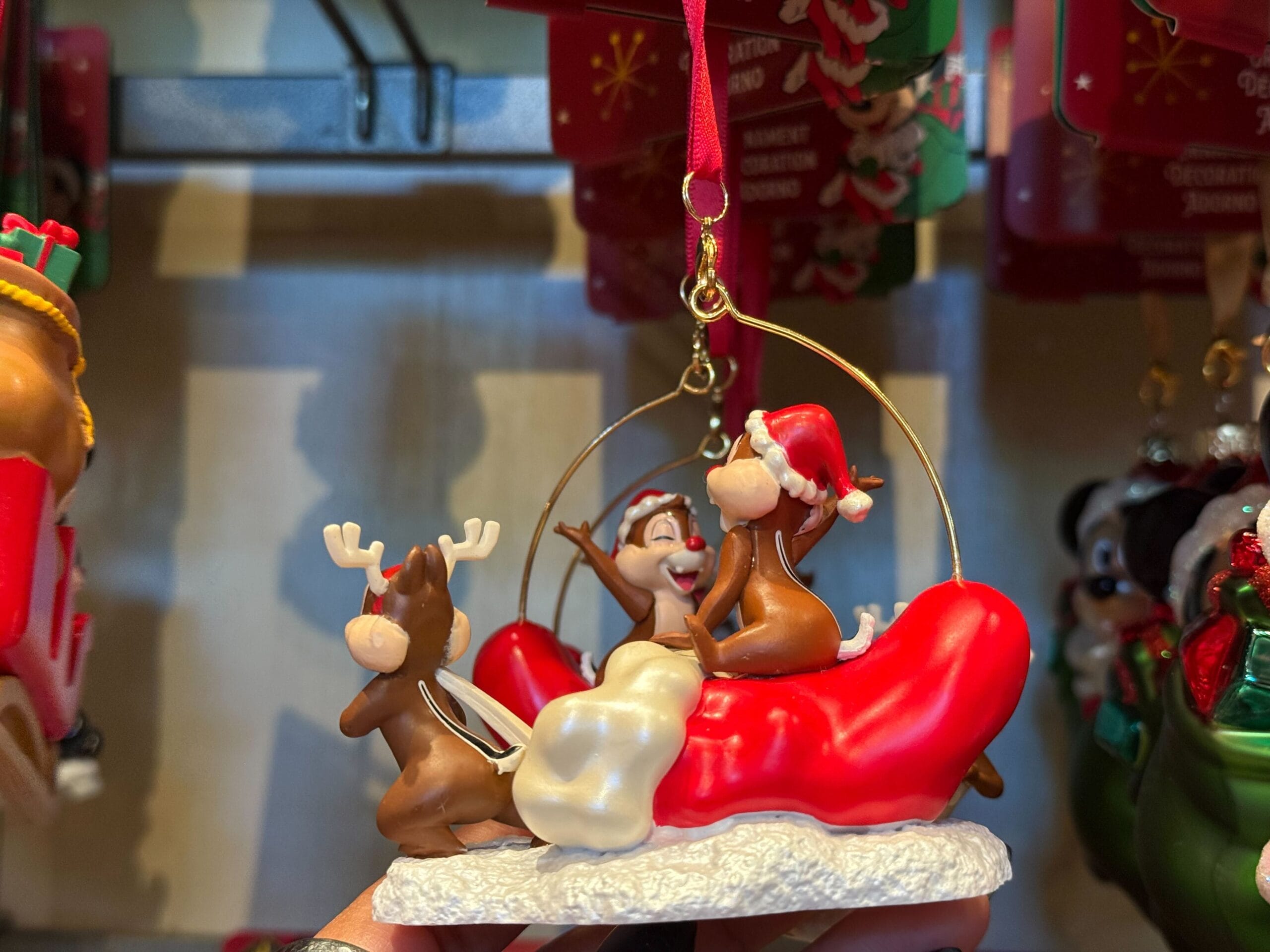 Three cartoon chipmunks in Santa hats ride a sleigh as a Disneyland Resort Christmas ornament, set against holiday park decor.