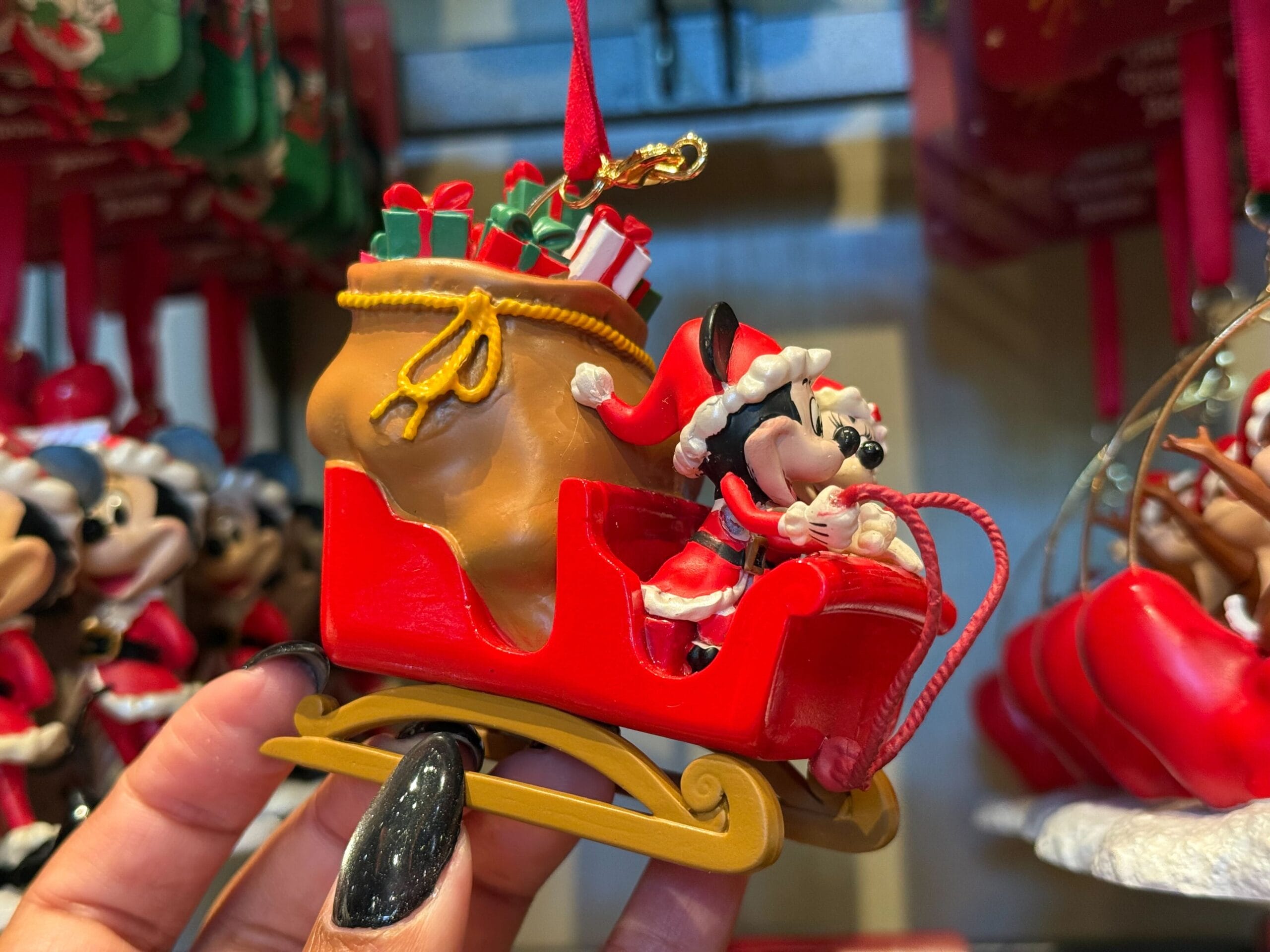A hand holds a Disney Parks Christmas ornament: Minnie Mouse as Santa in a red sleigh full of gifts, perfect for park fans.