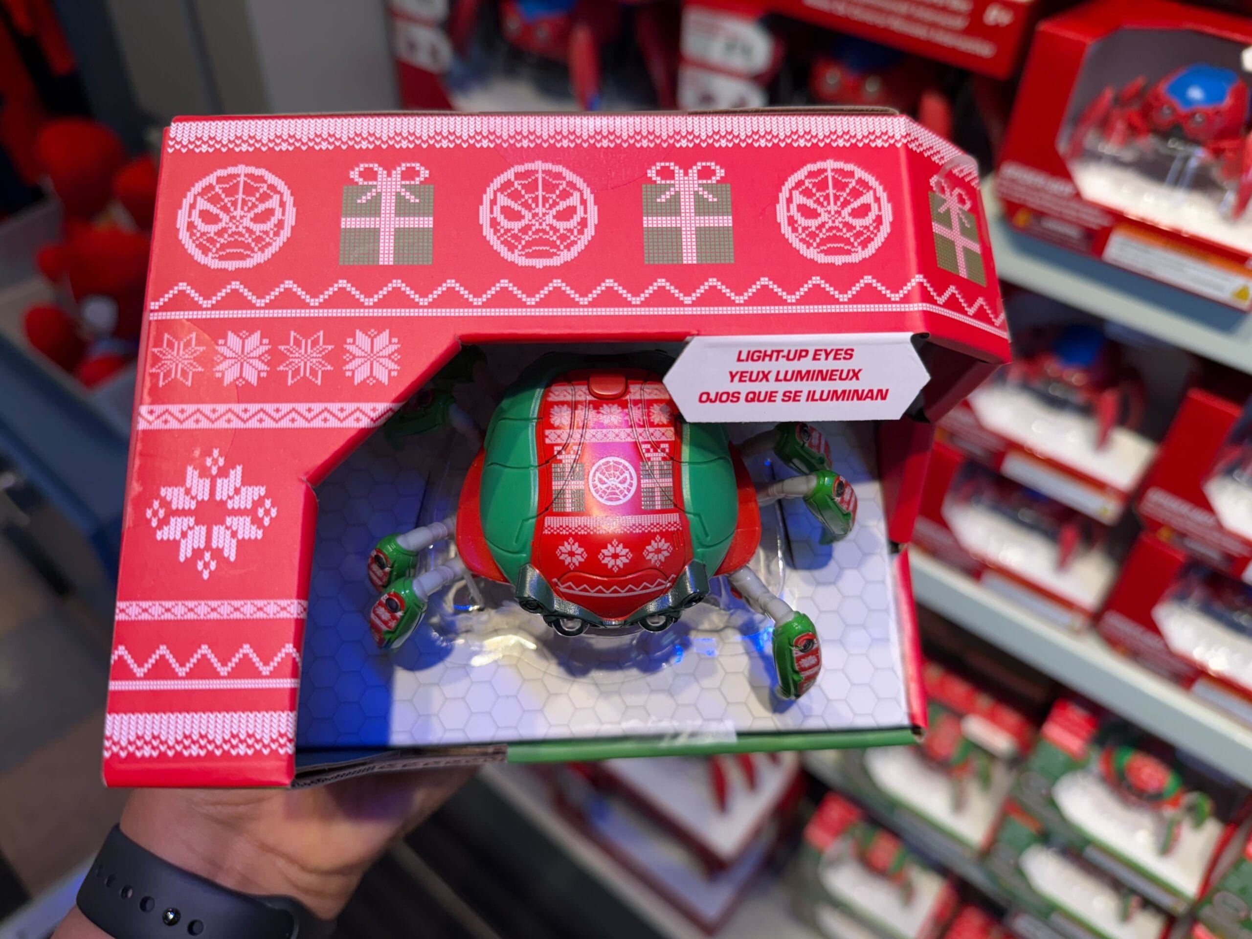 A hand displays a festive Spider-Bot in red holiday packaging at Disneyland, decorated with snowflakes and gifts, Main Street style.