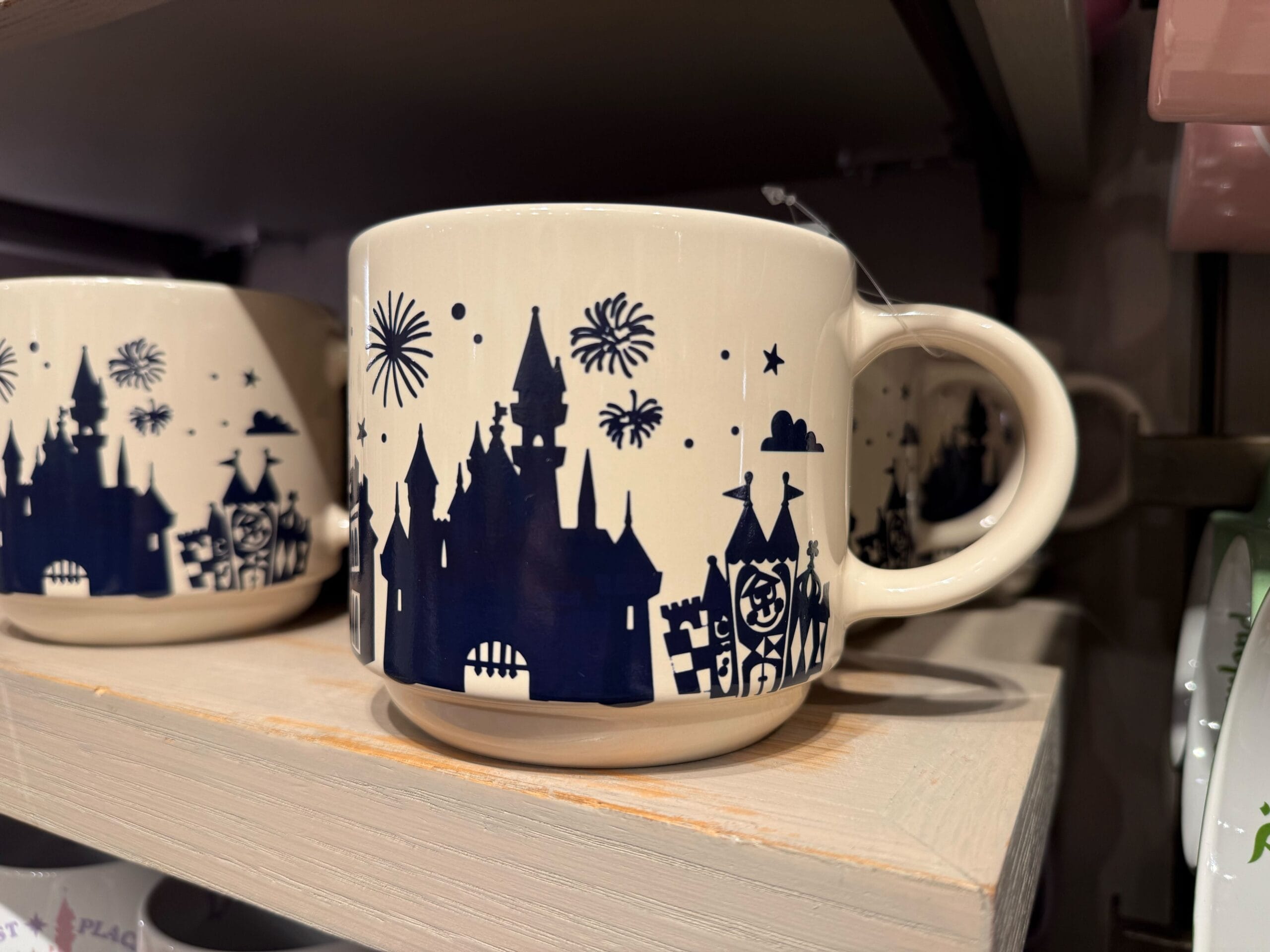 Cream mug featuring a navy blue castle reminiscent of Disneyland's Sleeping Beauty Castle, fireworks and stars, on a shelf with other mugs.