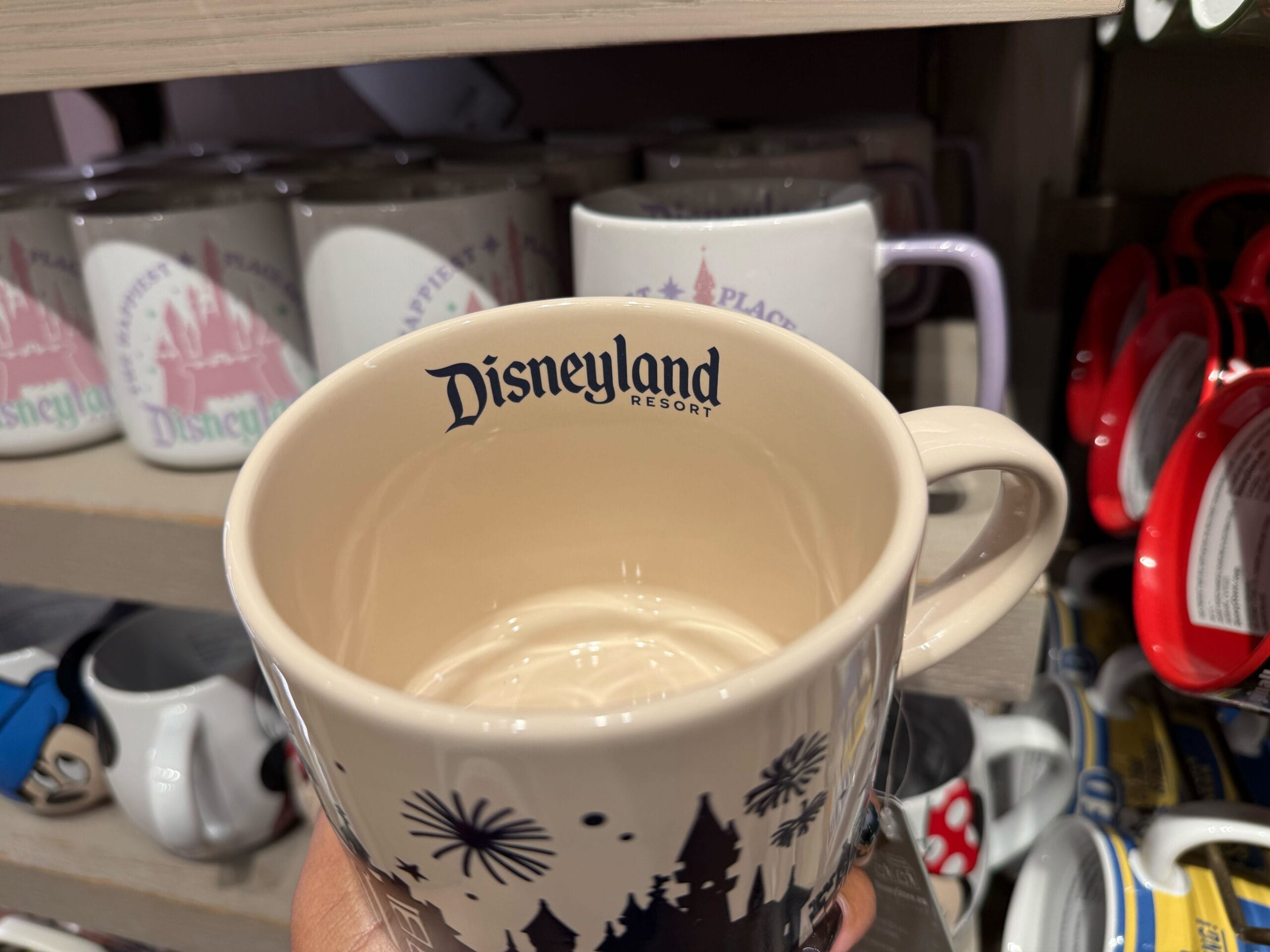 A hand displays a Disneyland Resort mug before shelves lined with Disney mugs, reminiscent of park gift shops near iconic castles.