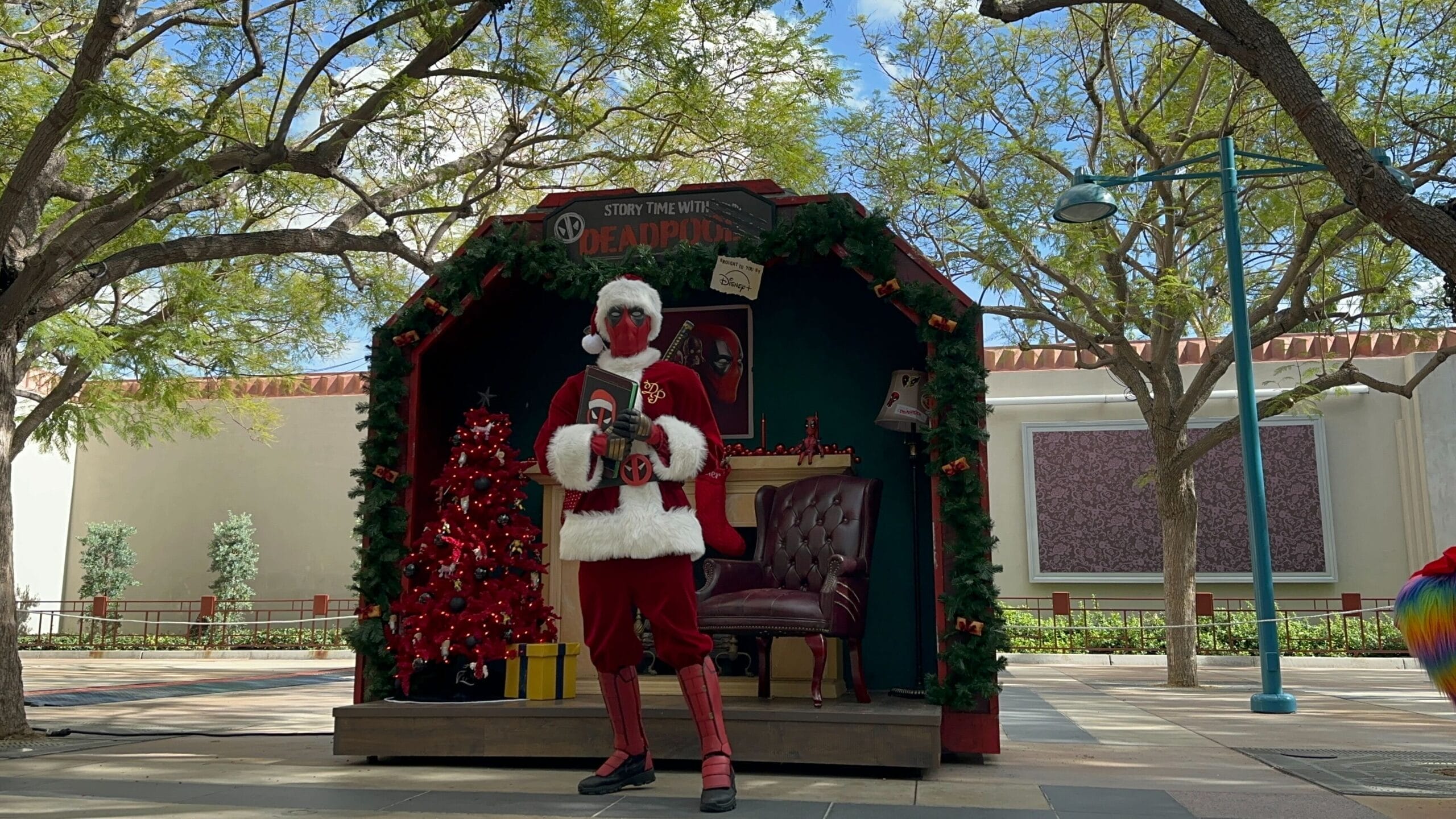 A person dressed as Deadpool in a Santa suit poses by a holiday display at Disneyland, with Sleeping Beauty Castle in the distance.