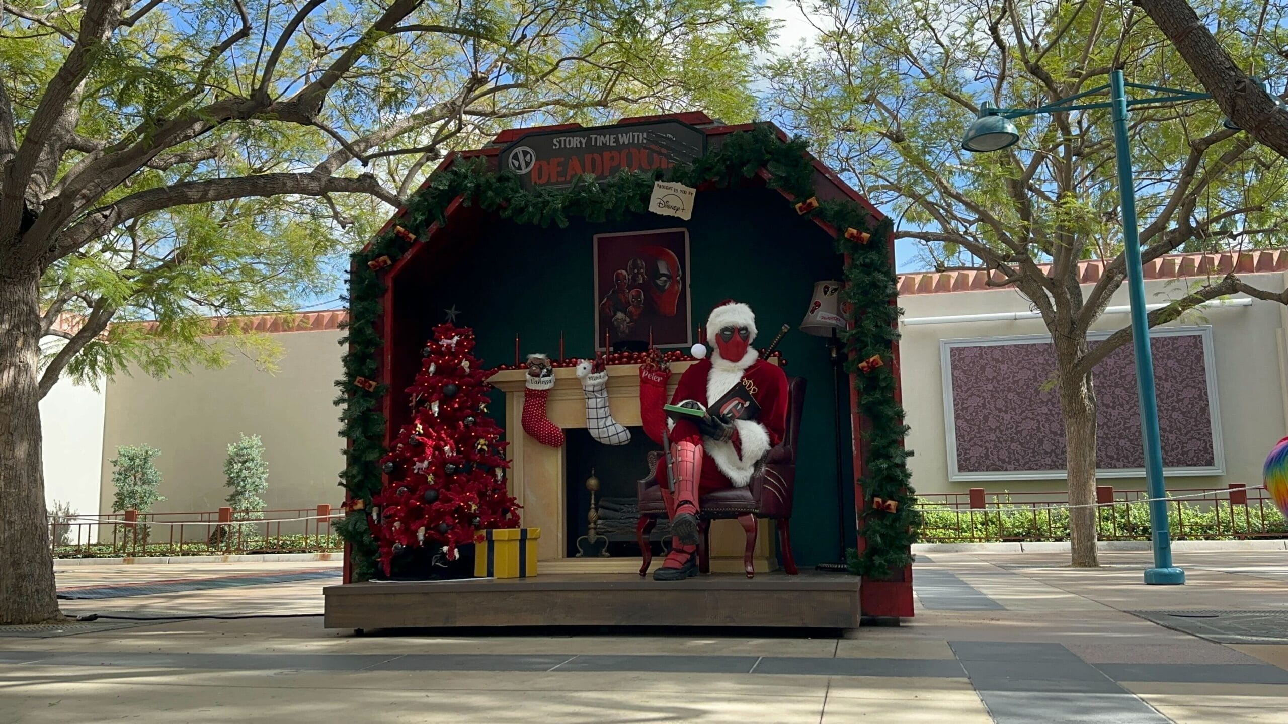 Deadpool in a Santa suit reads by a decorated fireplace at Disneyland Resort’s holiday display, Sleeping Beauty Castle twinkling nearby.