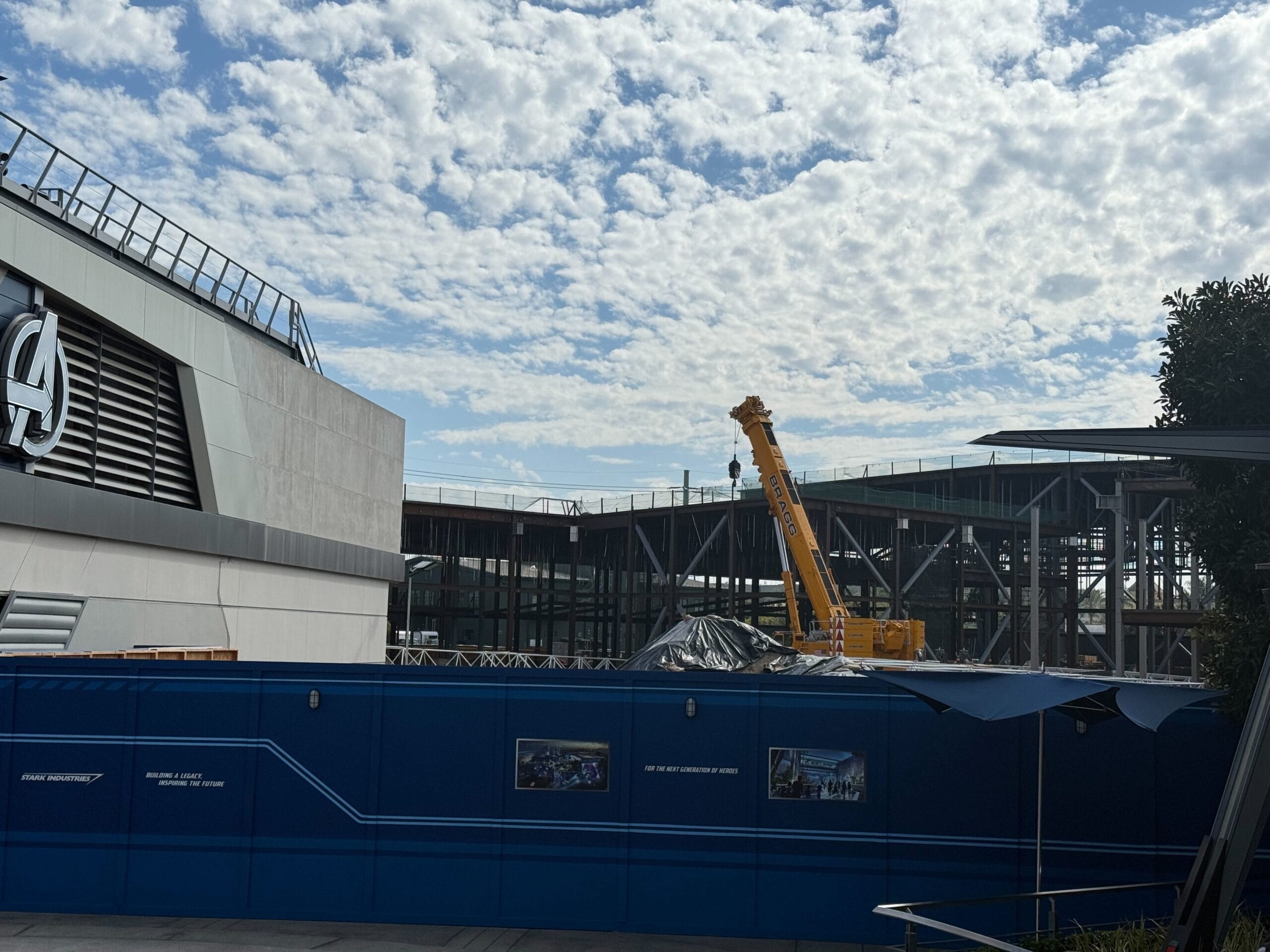 A construction site with a yellow crane behind a blue wall, near an Avengers-themed building like those at Disney California Adventure.