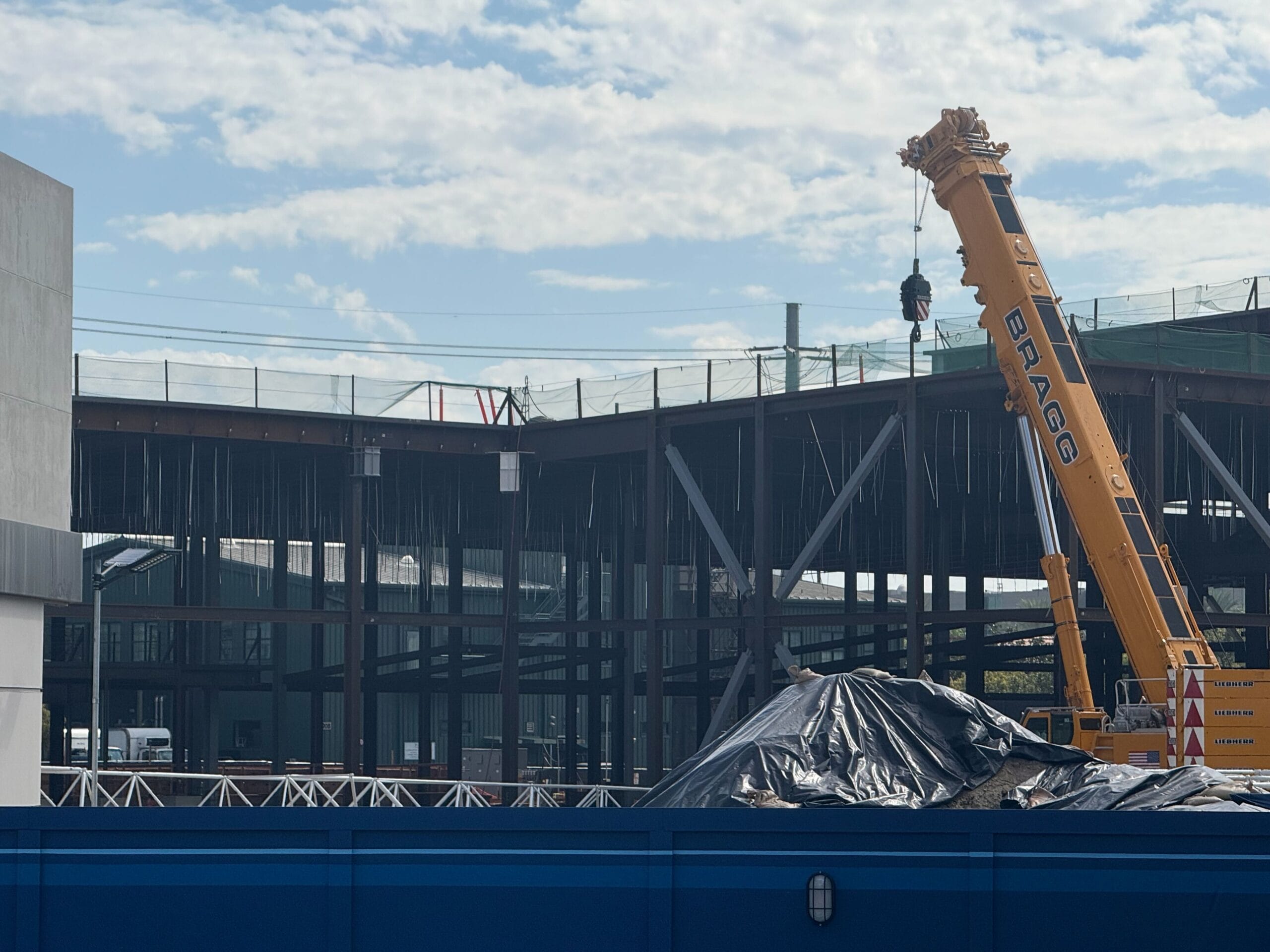 A yellow crane lifts materials at a steel-framed building site behind a blue barrier, typical of Disney park construction zones.