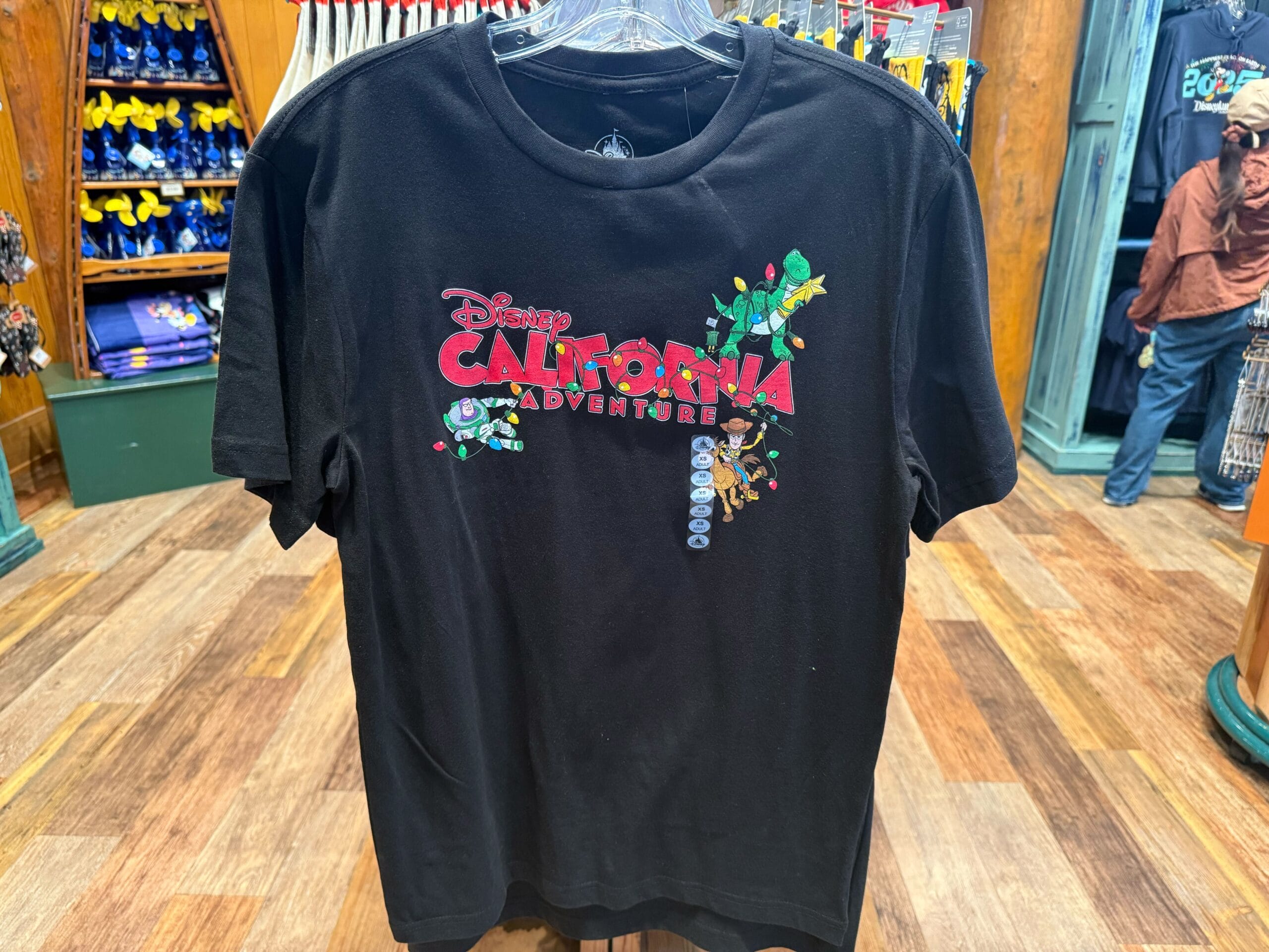 Black T-shirt with “Disney California Adventure” and vibrant Disney character graphics, displayed in a park merchandise store.