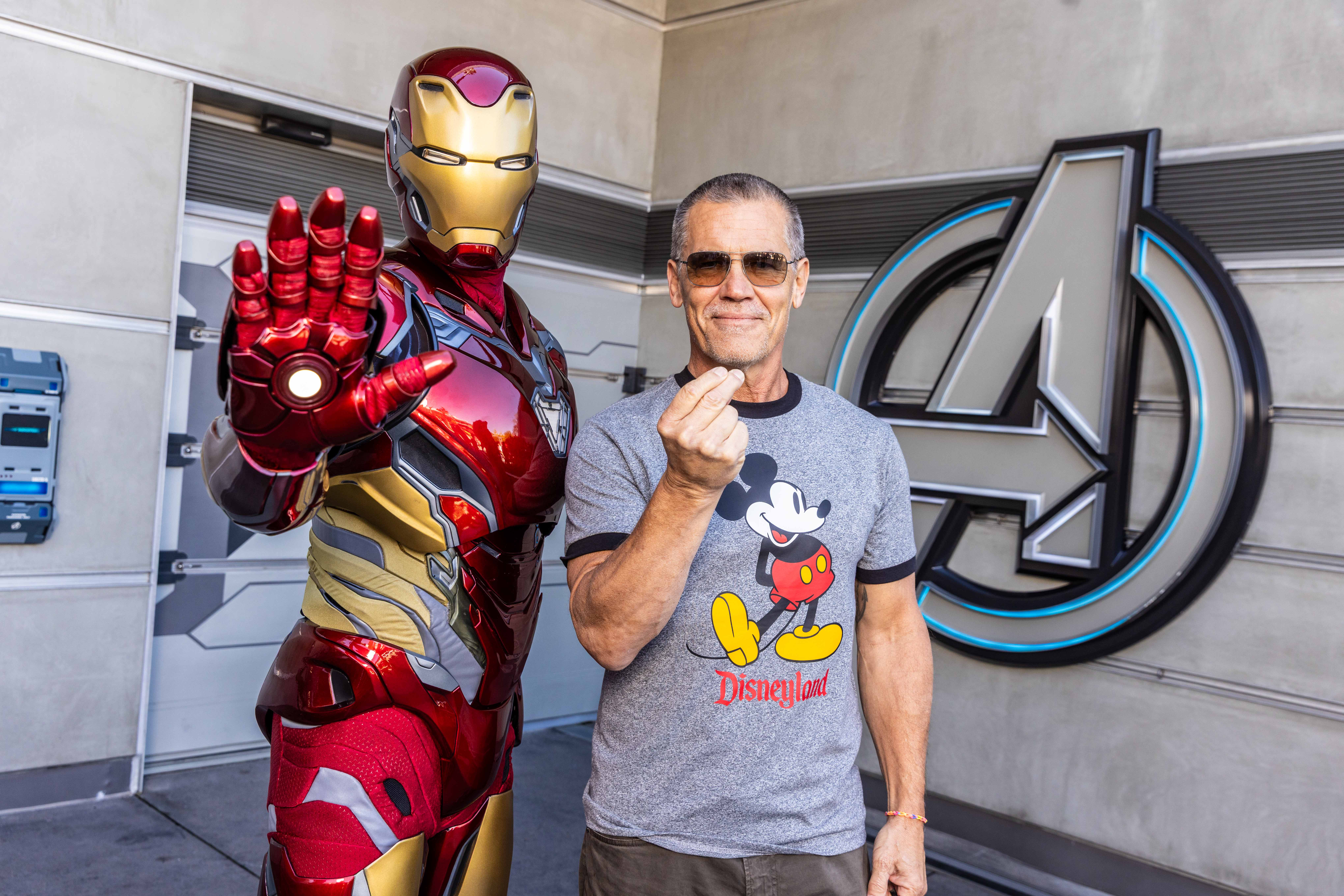 A man in a Mickey Mouse shirt strikes a pose with Iron Man at Avengers Campus, both gesturing, no castle like those in Disney parks.
