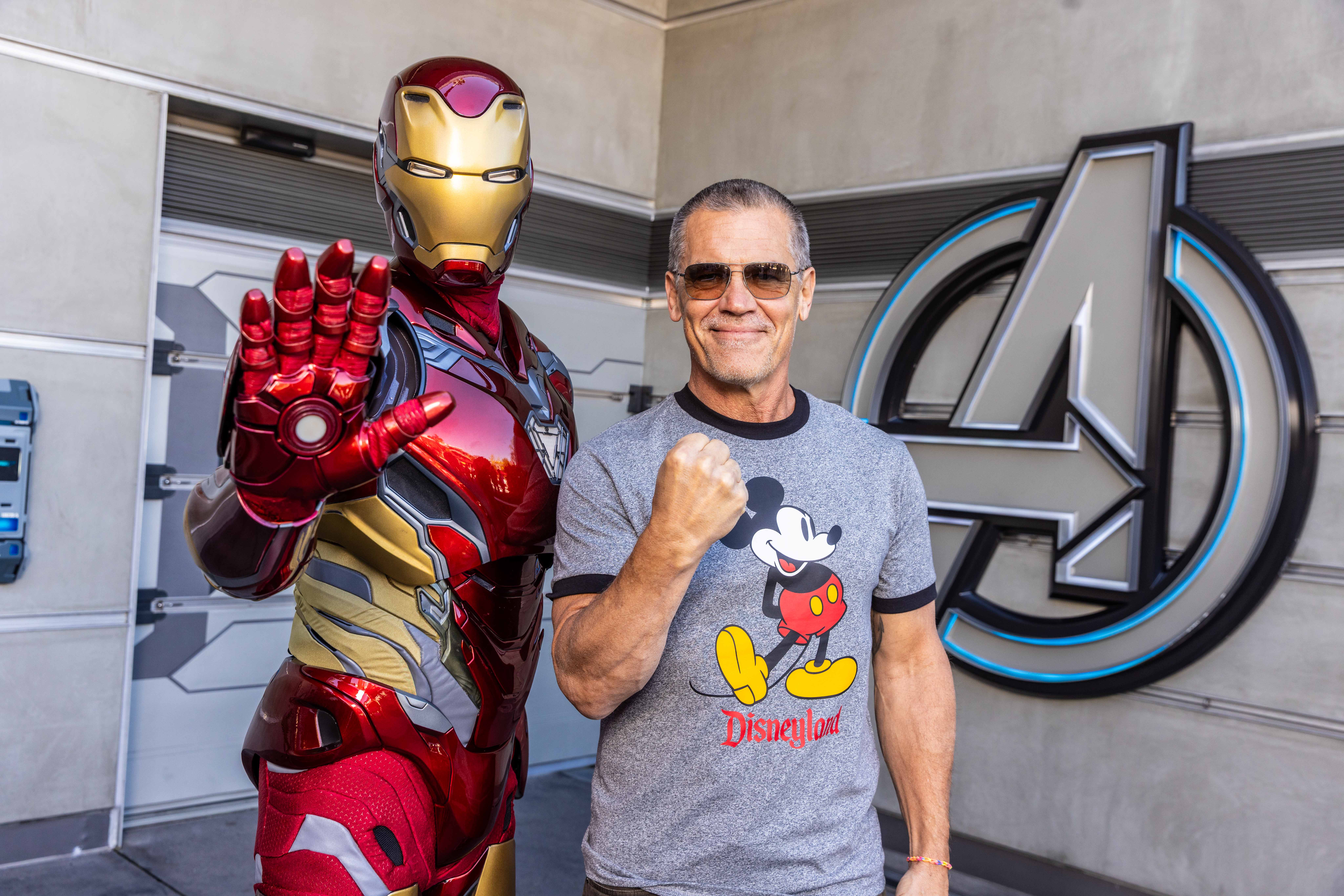 A man in a Disneyland shirt poses with Iron Man at Avengers Campus, both striking energetic poses near iconic Disney details.