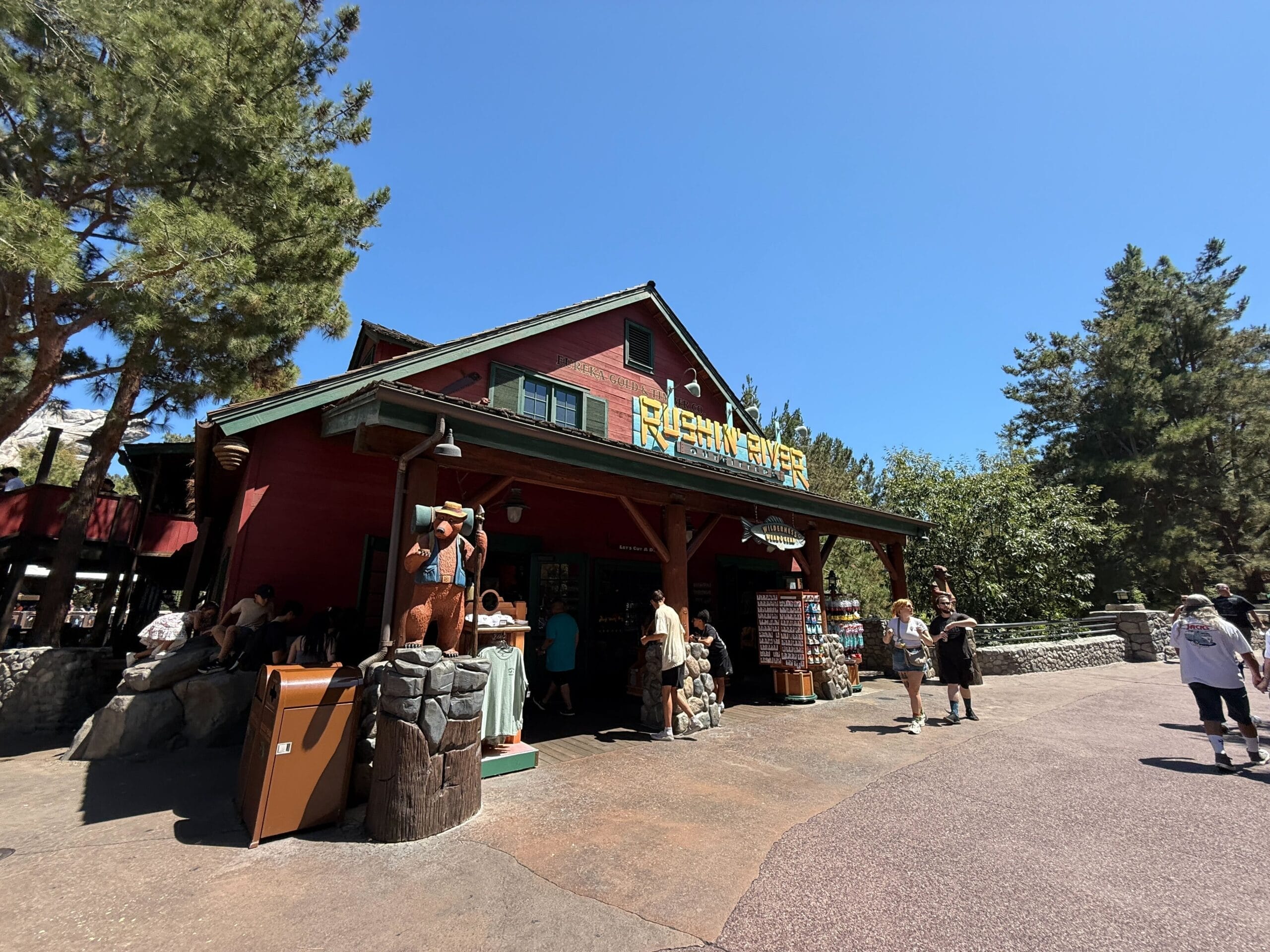 Guests gather outside the woodsy Rushin' River shop at Disney California Adventure, surrounded by trees under a bright sky.