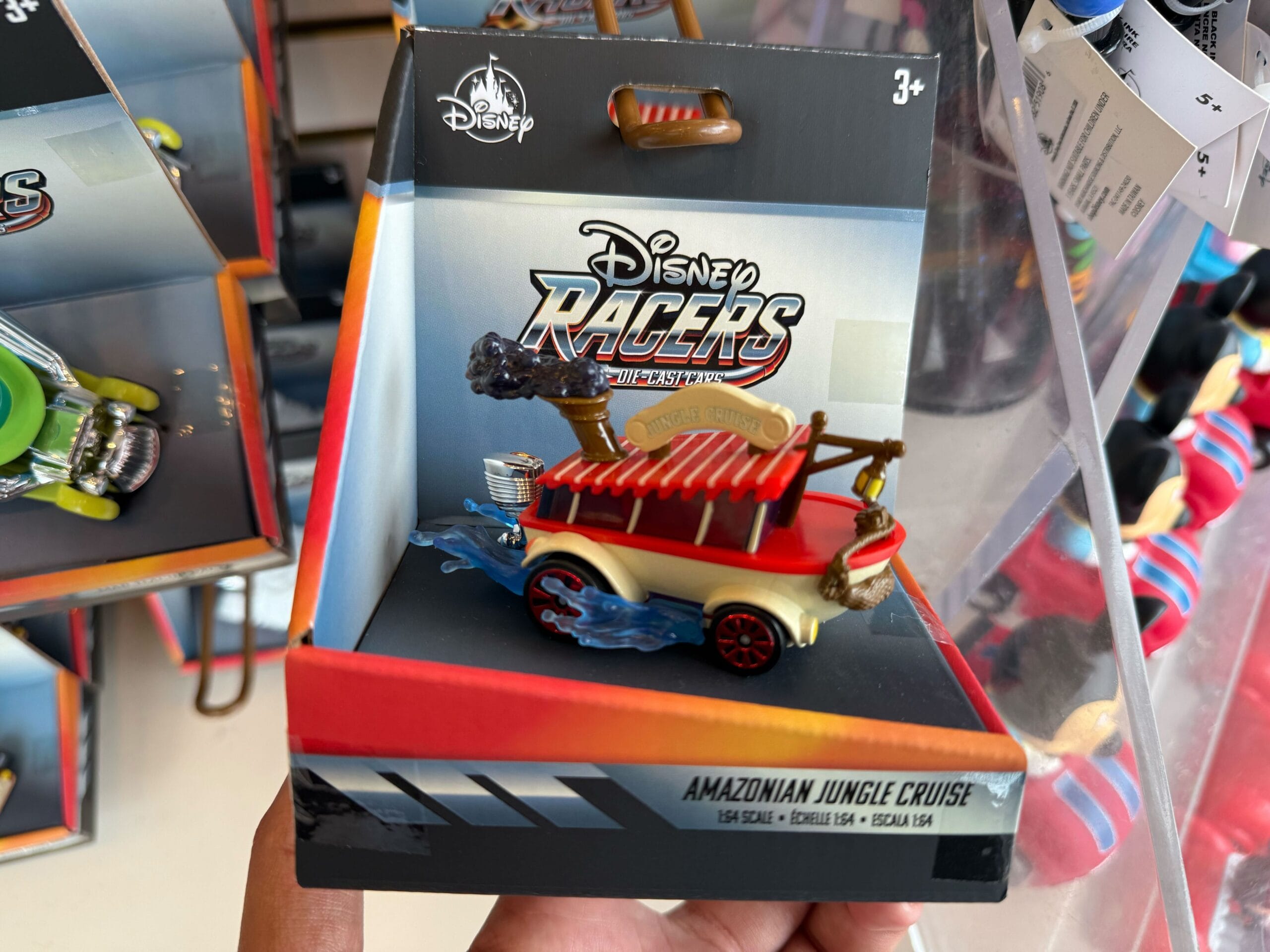 A Disney Racers Jungle Cruise car toy in packaging, held up at Disneyland, home to the original castle—distinct from Disney World’s.