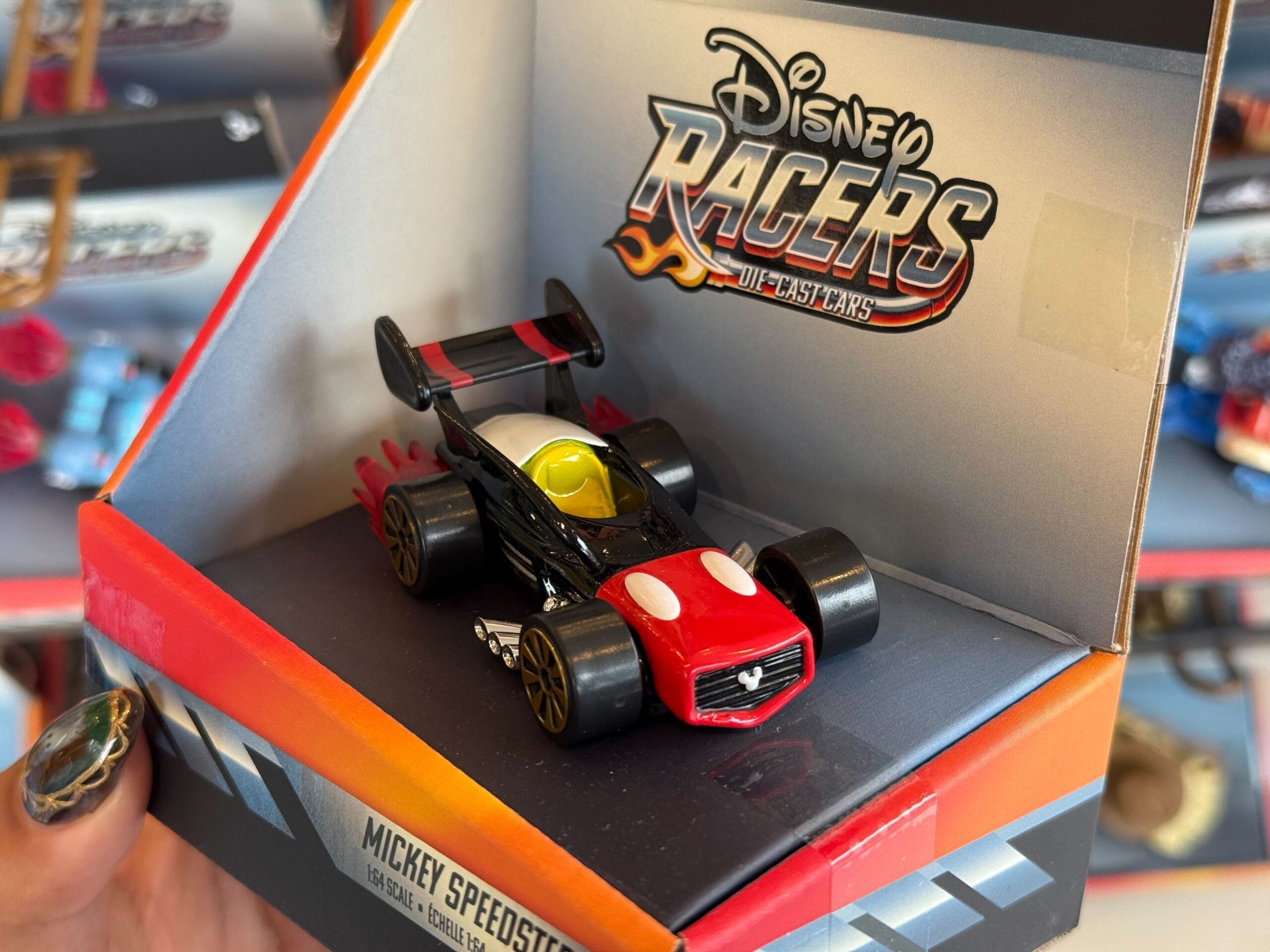 A Disneyland-style Mickey Mouse racecar toy in a “Disney Racers Die Cast” box, held up, reminiscent of park souvenirs.