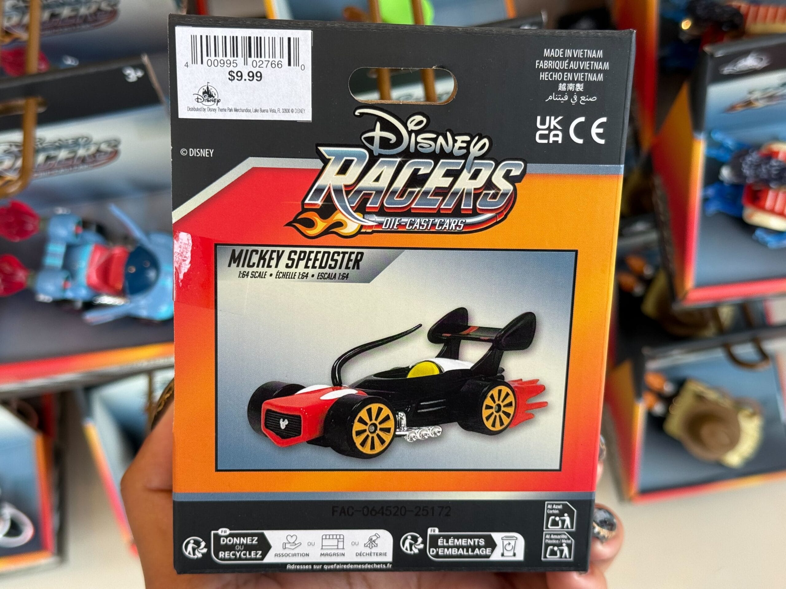 A hand holds a Disney Racers Mickey Speedster car package, displayed in a Disneyland shop; castle not pictured for comparison.