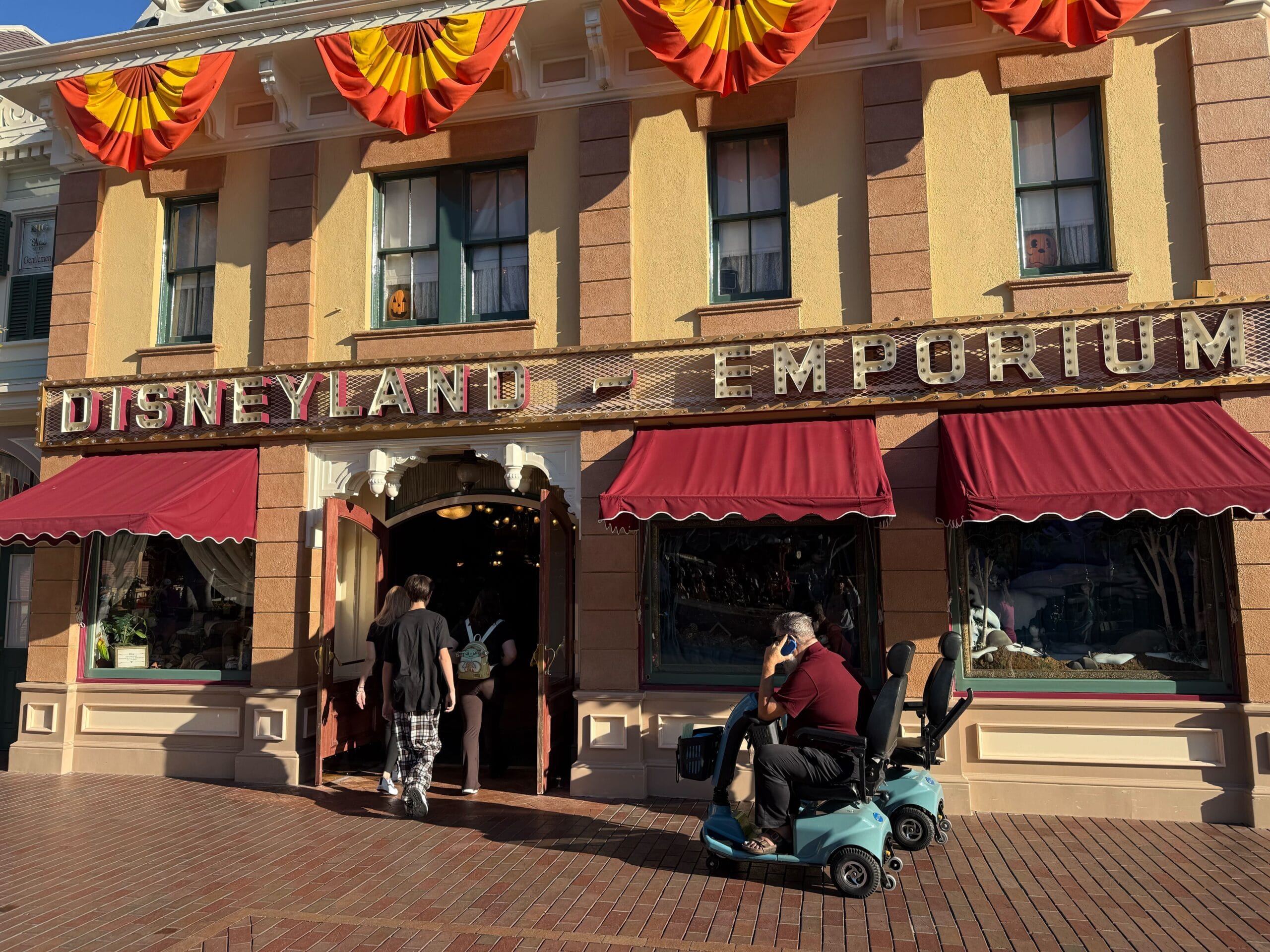 People enter Disneyland Emporium under red awnings; someone in a Logo Sweatshirt is near the store entrance.