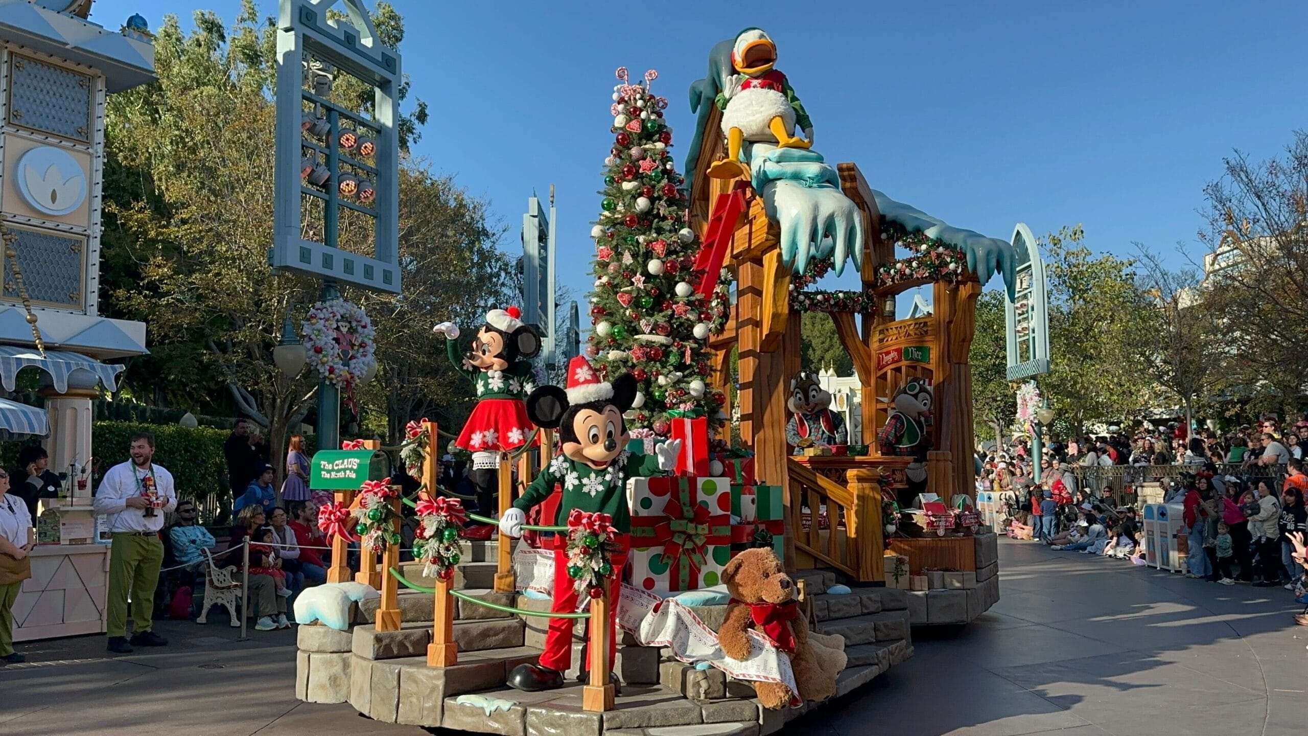 A cheerful Disney holiday parade float with Mickey, Minnie, and Donald by Sleeping Beauty Castle, smaller than Florida’s Cinderella Castle.
