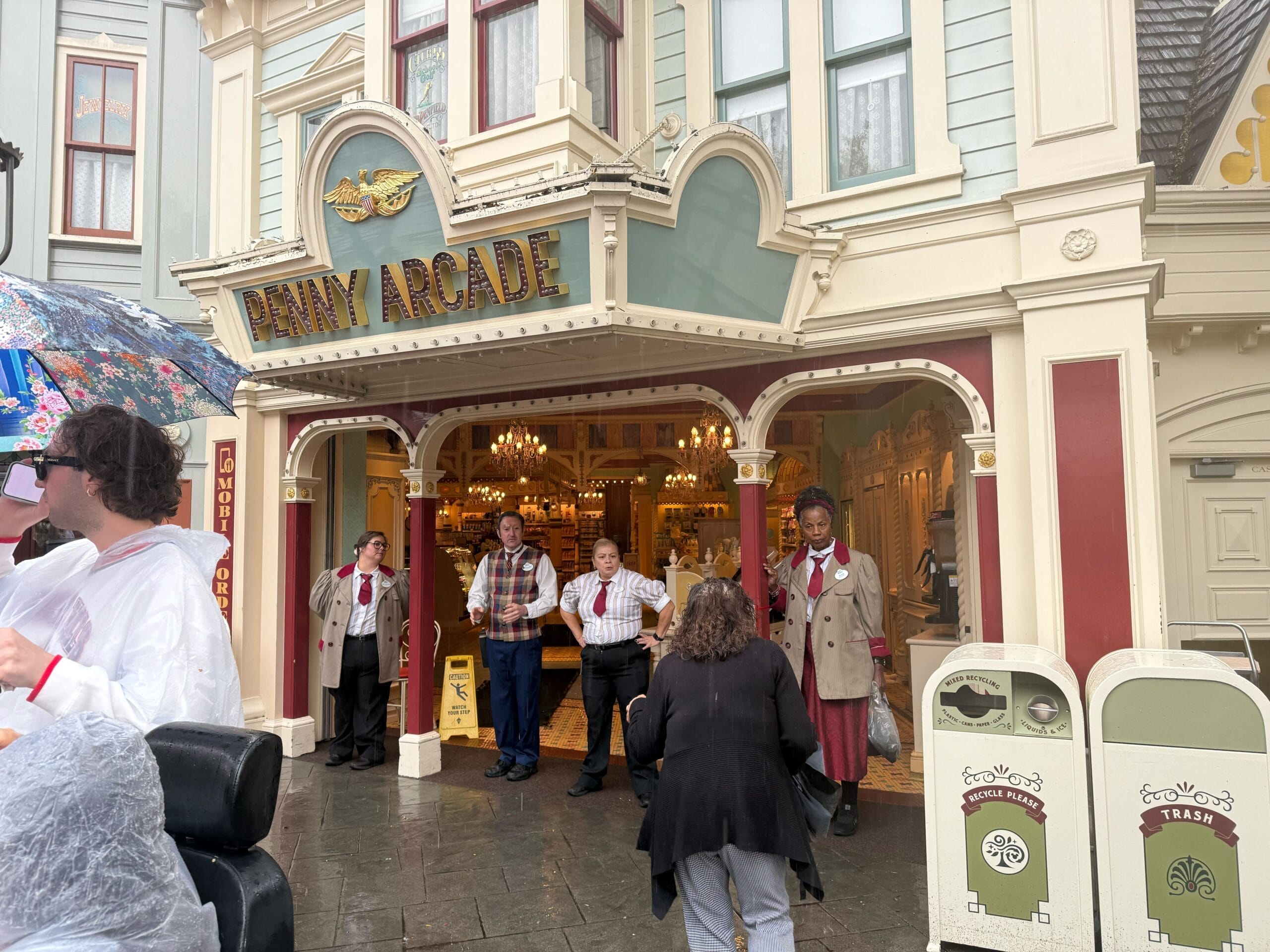 Guests in rain ponchos and wait outside Disneyland’s Main Street Penny Arcade during an evacuation as four cast members block the entrance.