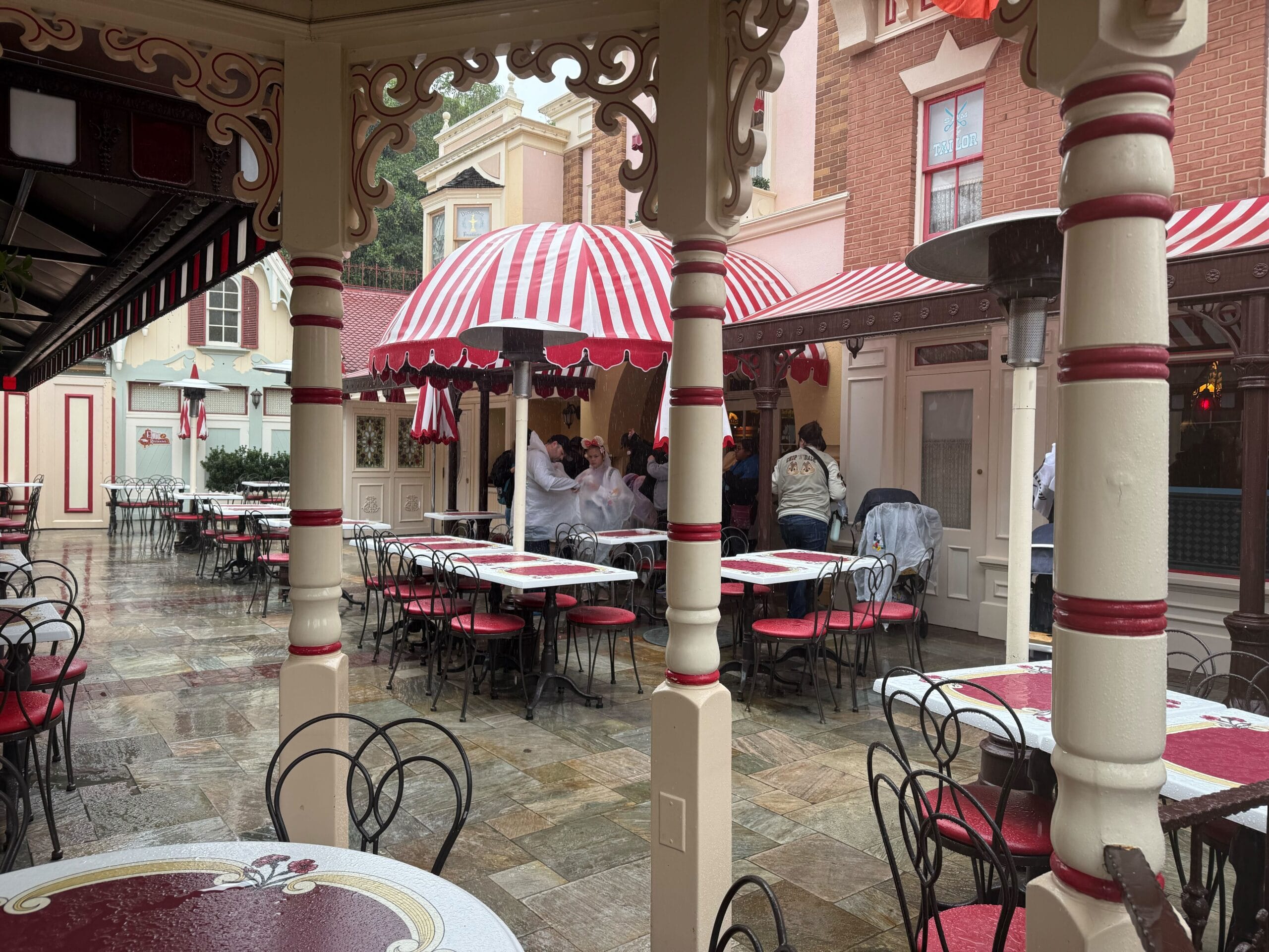 Outdoor café at a Disney park with red and white umbrellas, empty tables, and guests taking cover from rain near the entrance.