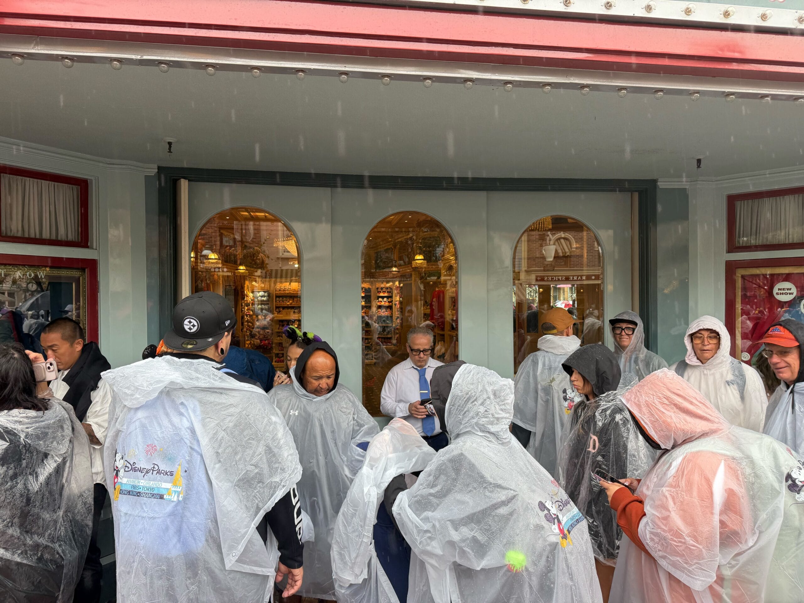 A group in Disney rain ponchos stands outside a park shop on a rainy day, some checking their phones, Main Street visible nearby.
