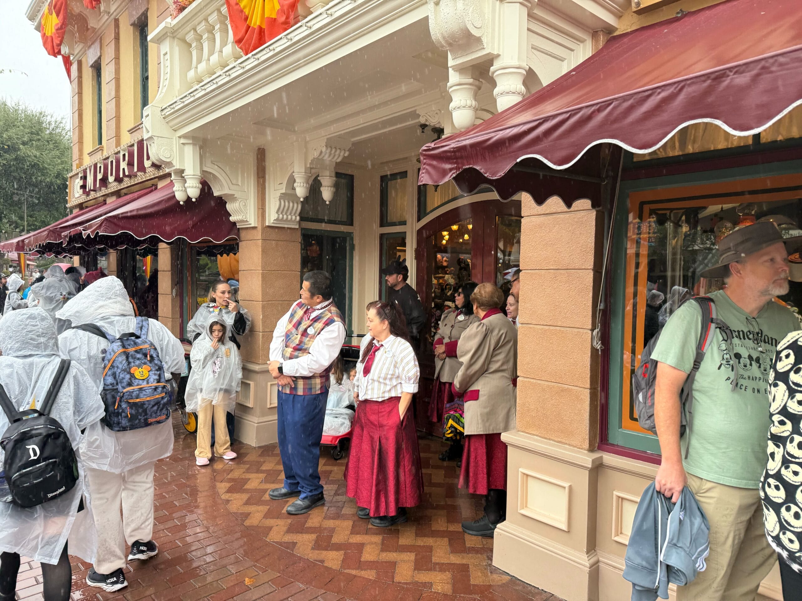 Guests in ponchos and cast members in themed attire stand outside a shop on a rainy day at a Disney park.
