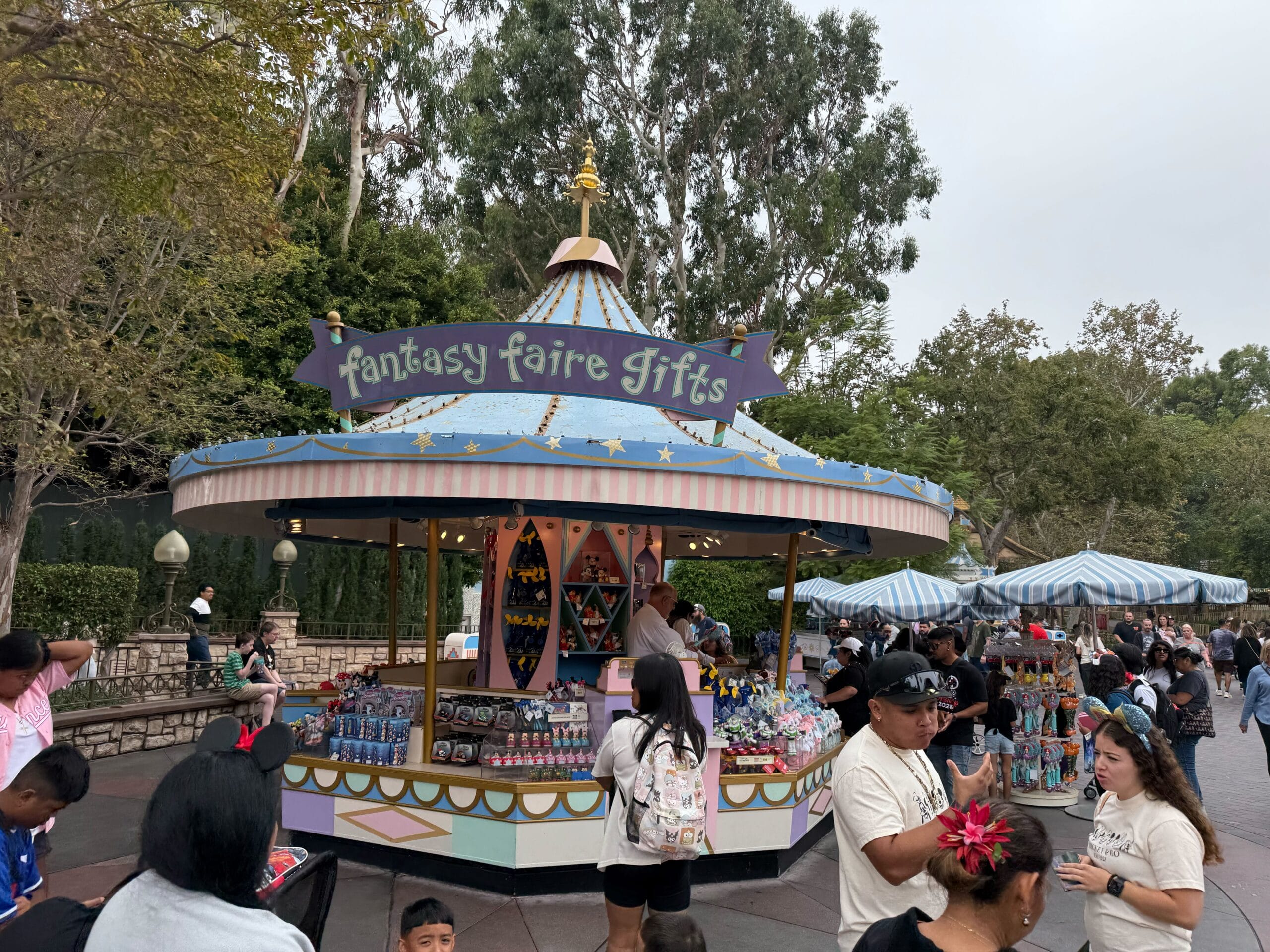 A vibrant Fantasy Faire Gifts kiosk at Disneyland offers Disney Racers toys, bustling with guests near Sleeping Beauty Castle.