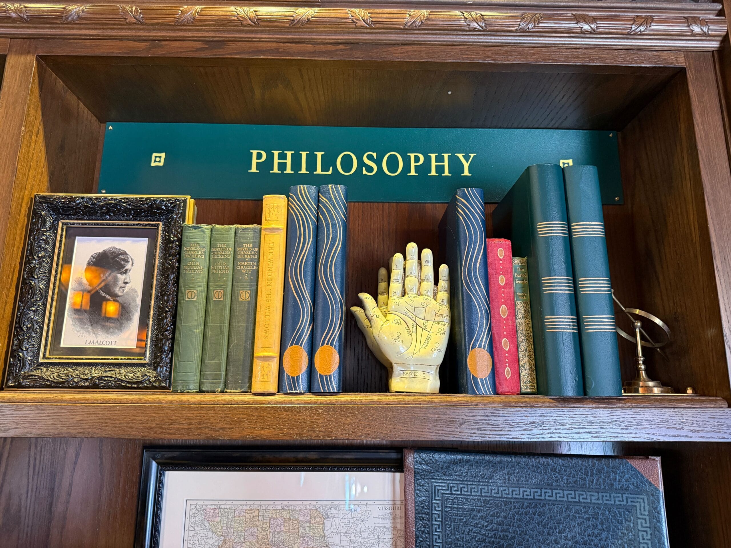 Books and decor on a wooden "Philosophy" shelf, including a framed photo, palmistry hand, and globe—reminiscent of Main Street’s vintage study displays in Walt Disney parks.