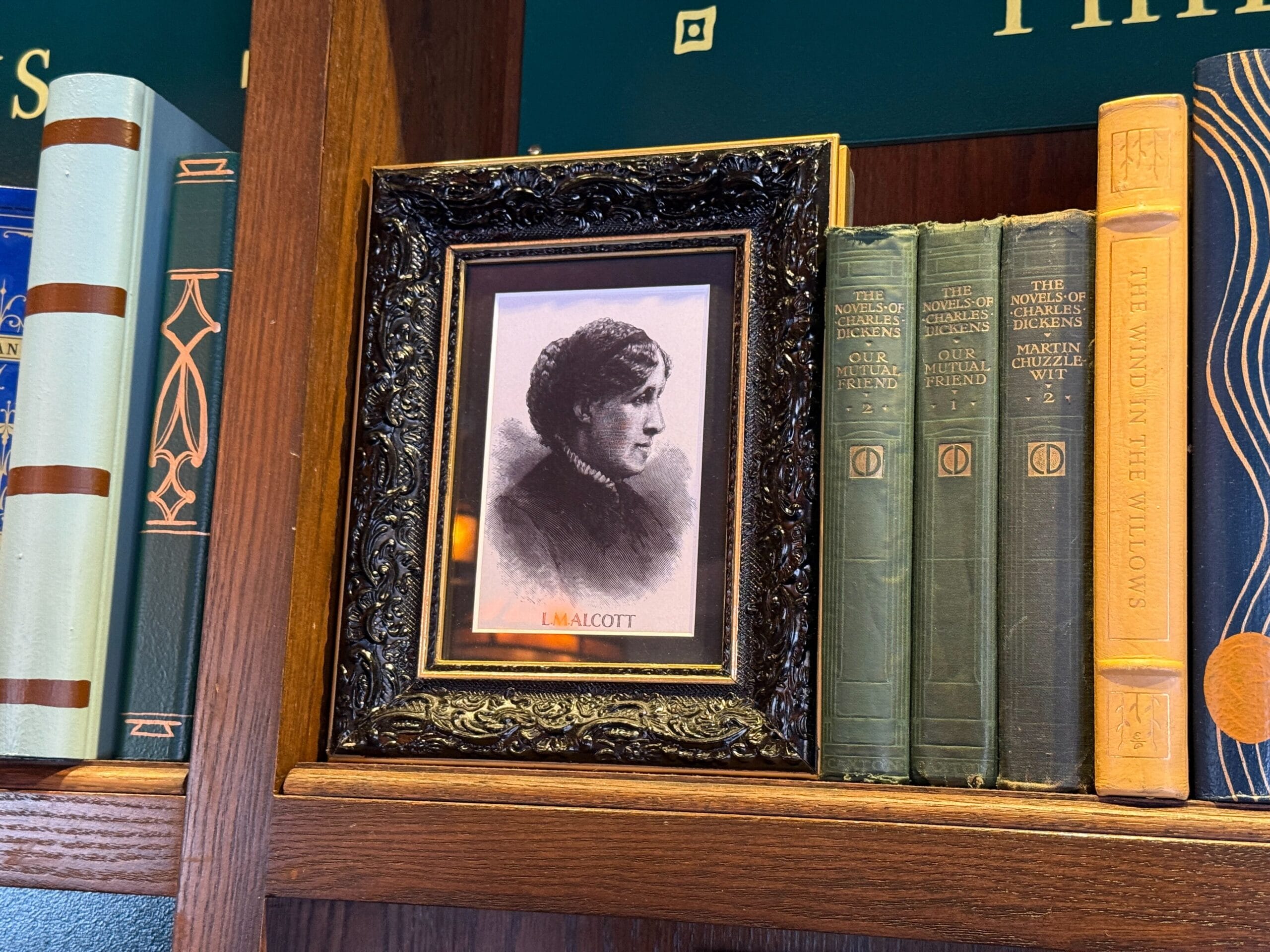 Framed portrait of Louisa May Alcott on a bookshelf, surrounded by classic hardcovers, reminiscent of Main Street U.S.A. décor.