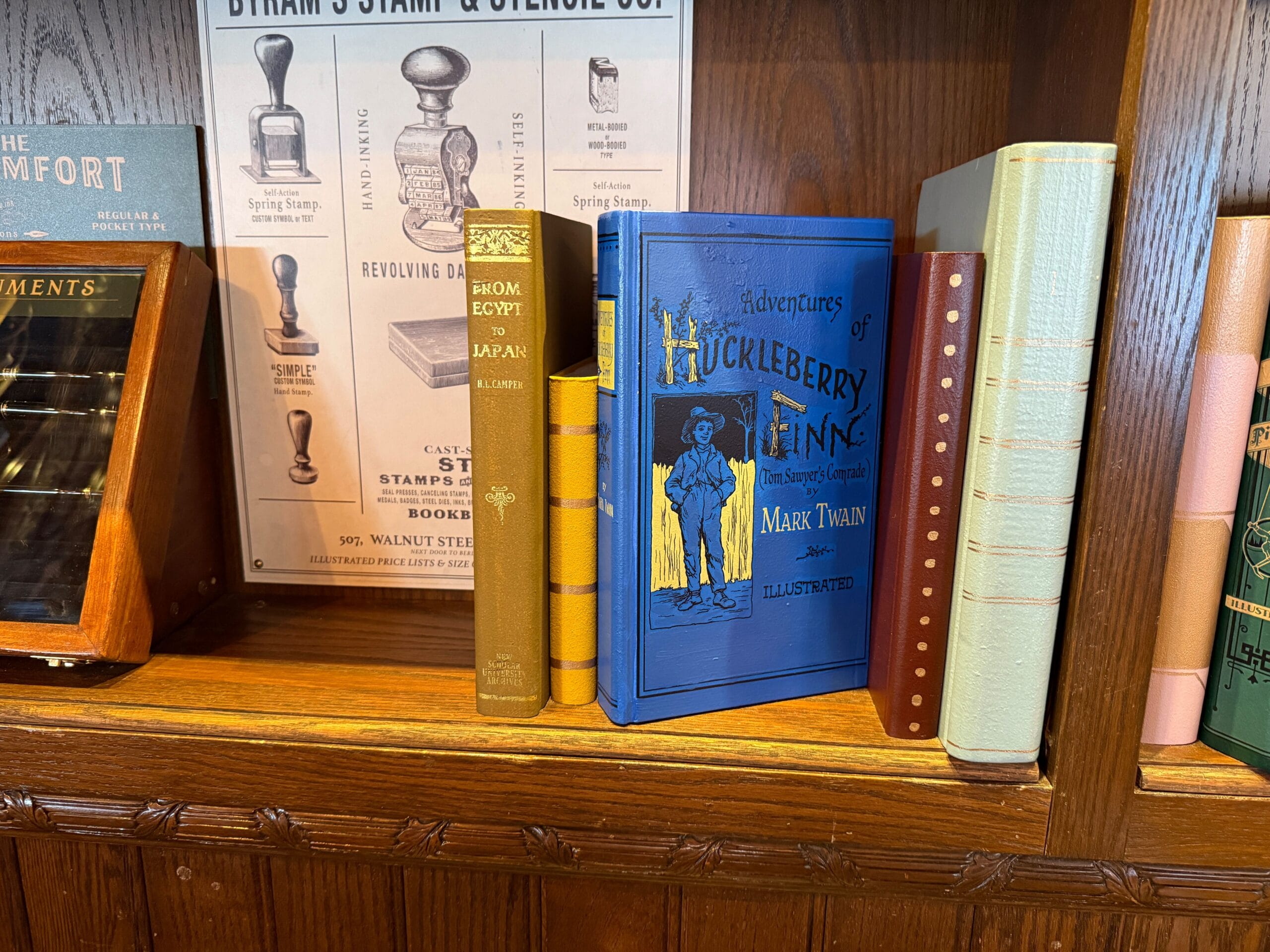 A blue "Adventures of Huckleberry Finn" book rests on a wooden shelf with vintage books, like those in Disneyland’s Emporium.