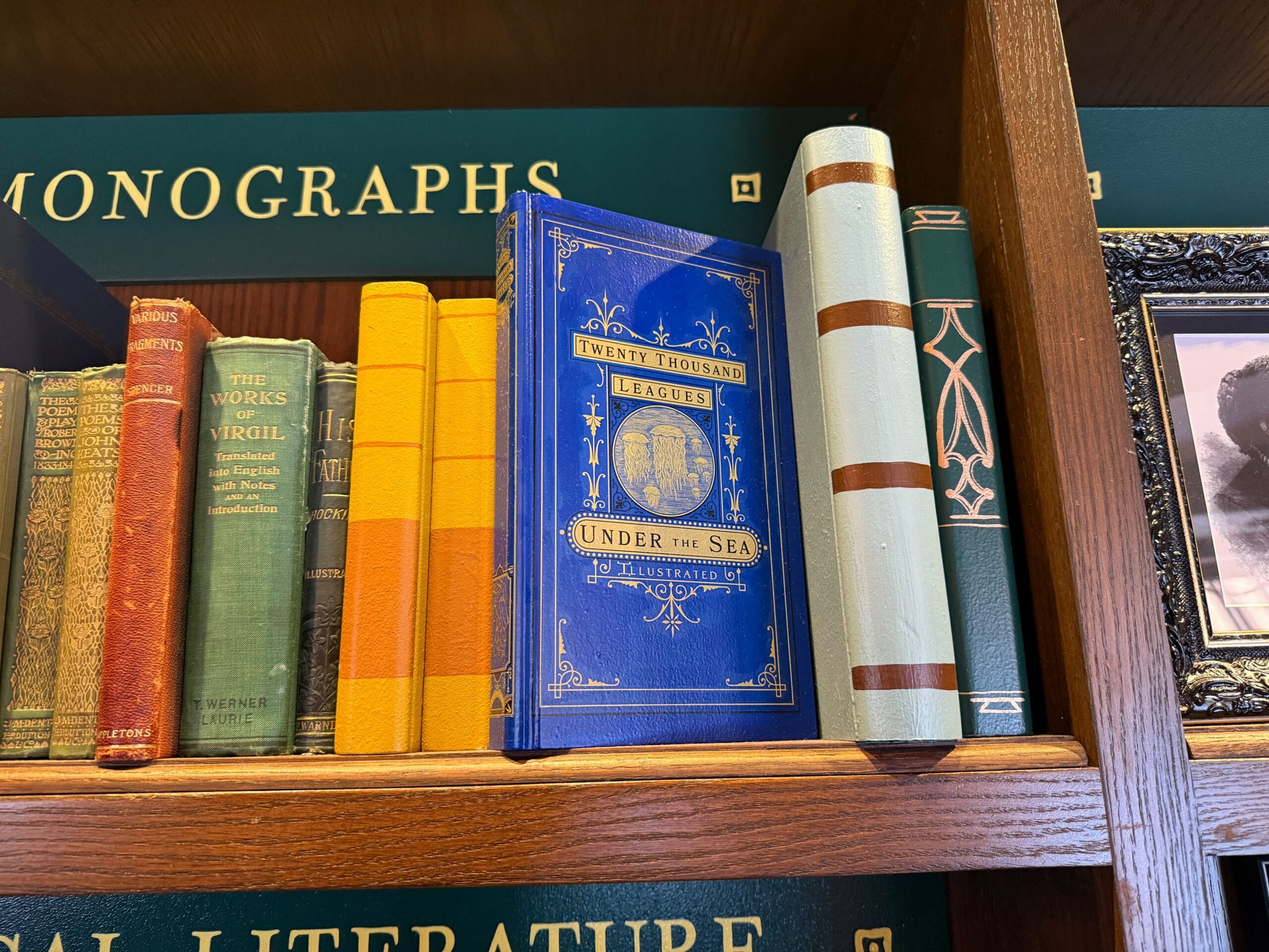 A blue "Twenty Thousand Leagues Under the Sea" book rests on a wooden shelf with vintage books, evoking classic Disney park decor.