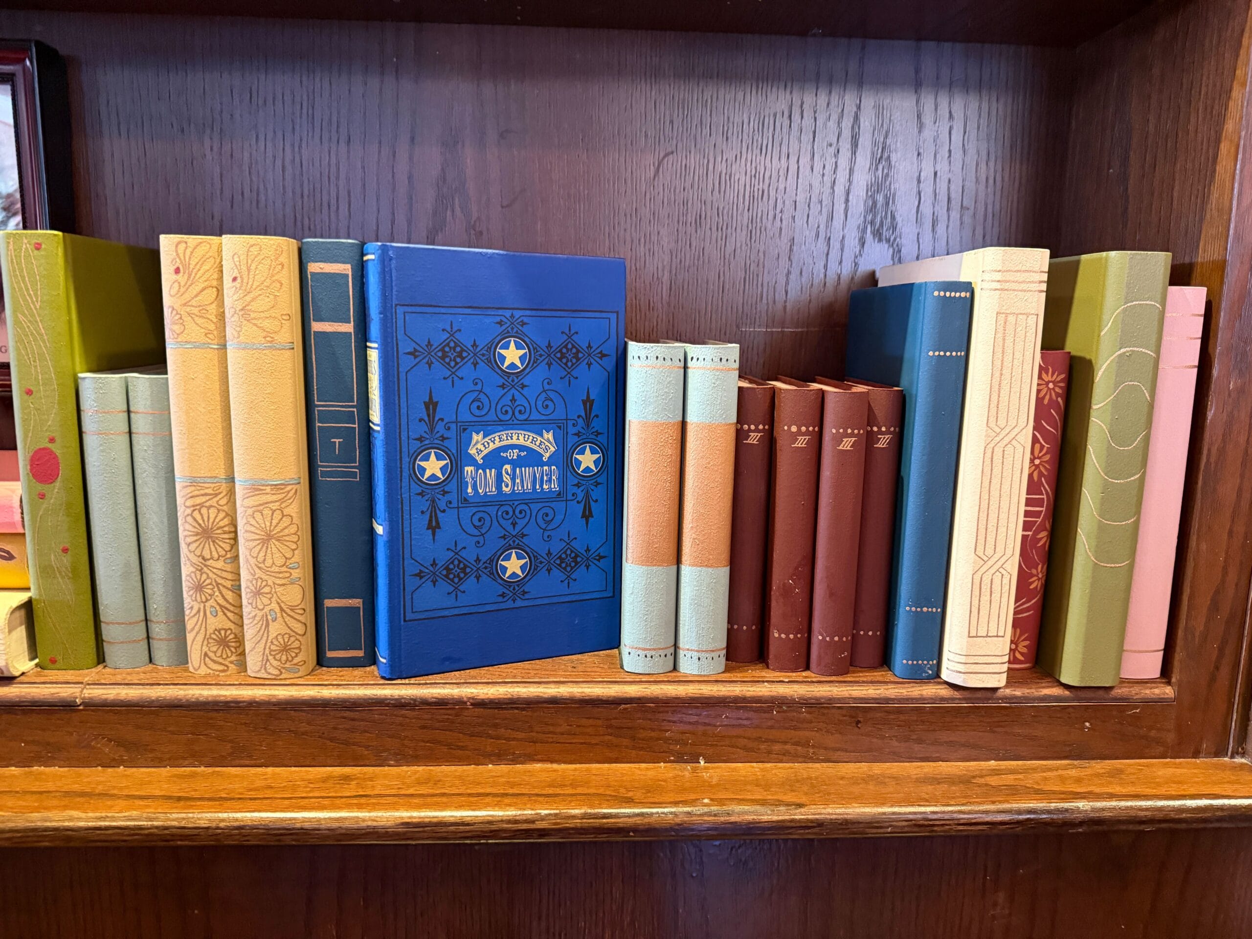A shelf of colorful vintage books, including a blue "The Adventures of Tom Sawyer," reminiscent of Main Street U.S.A. shops.